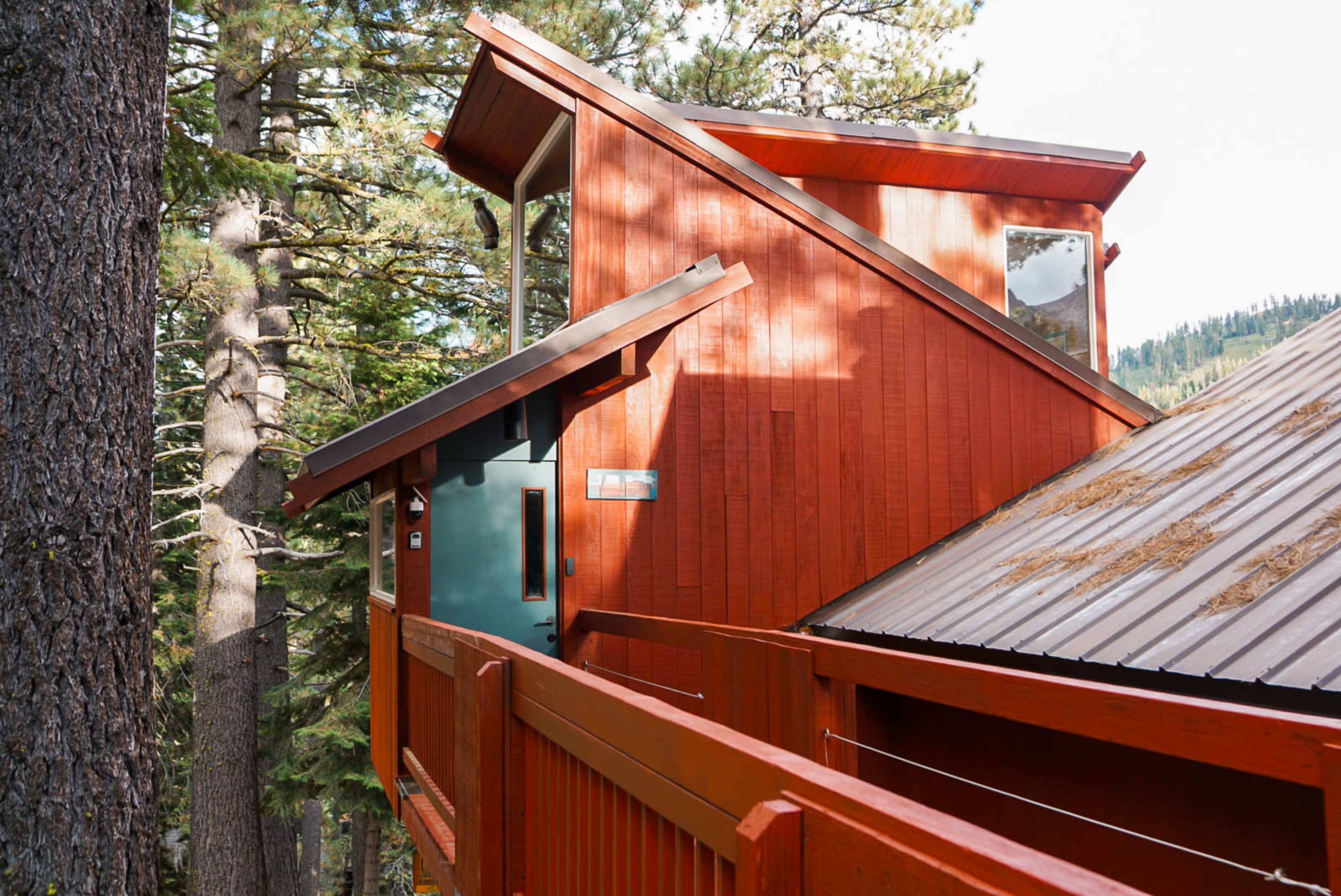 The image shows a modern, angular red cabin nestled among tall trees in a forested area.