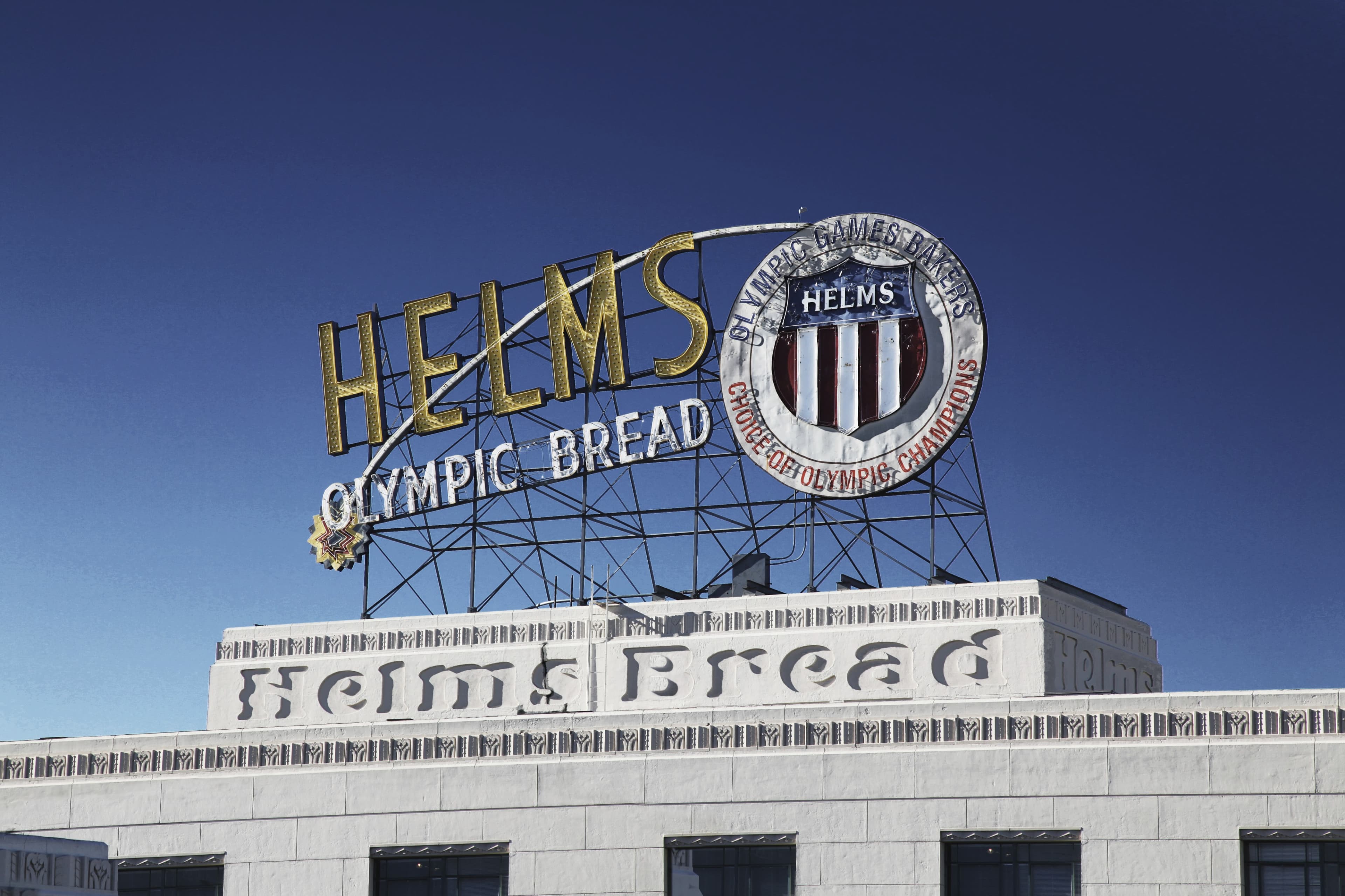The image features a large, vintage sign for Helms Bakery, prominently displaying "HELMS OLYMPIC BREAD" against a blue sky.
