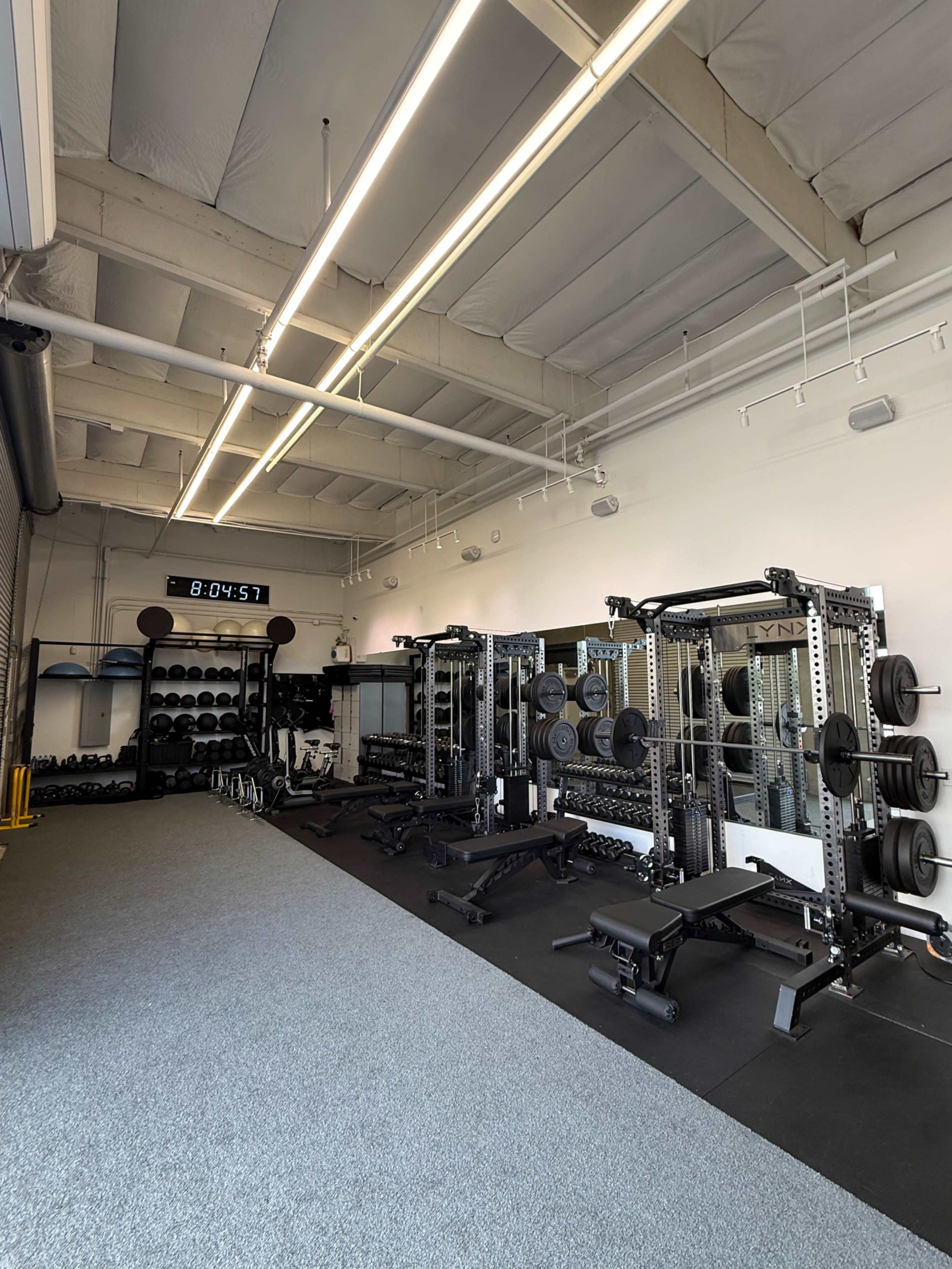 The image shows an interior view of a gym with weightlifting equipment, including racks, benches, and free weights arranged neatly along the walls.