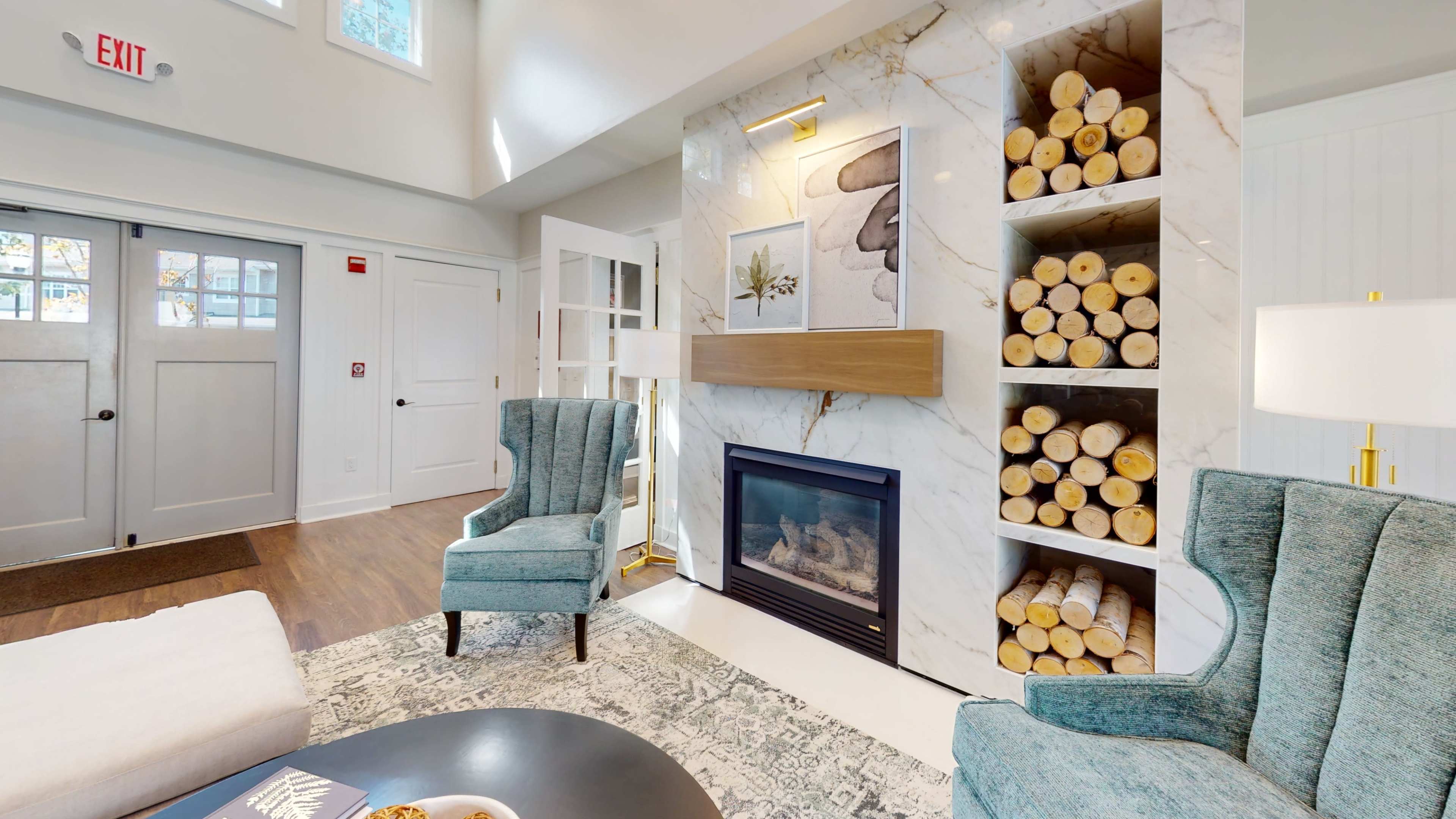 A cozy living room features a fireplace surrounded by stacked logs, two teal armchairs, and a decorative coffee table.
