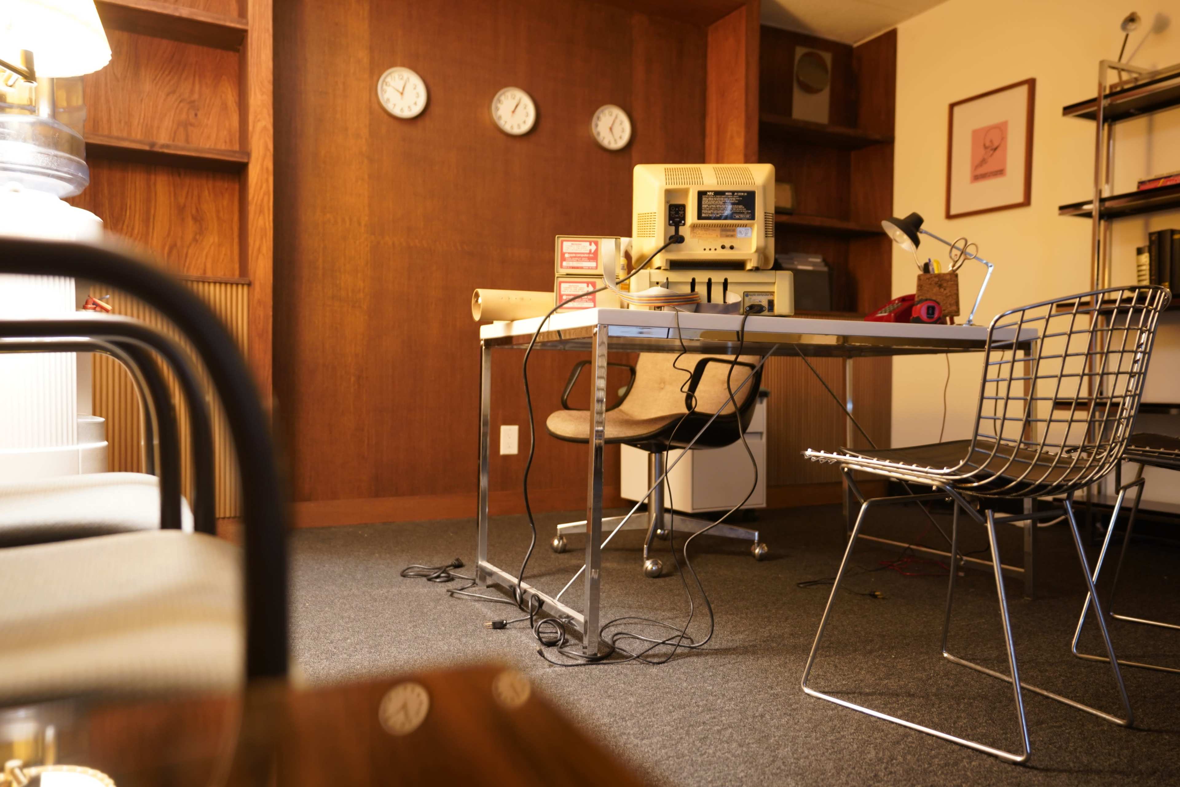 The image shows a minimalist office space with a wooden wall, clocks, a desk with an old computer and microphone, and a couple of chairs.