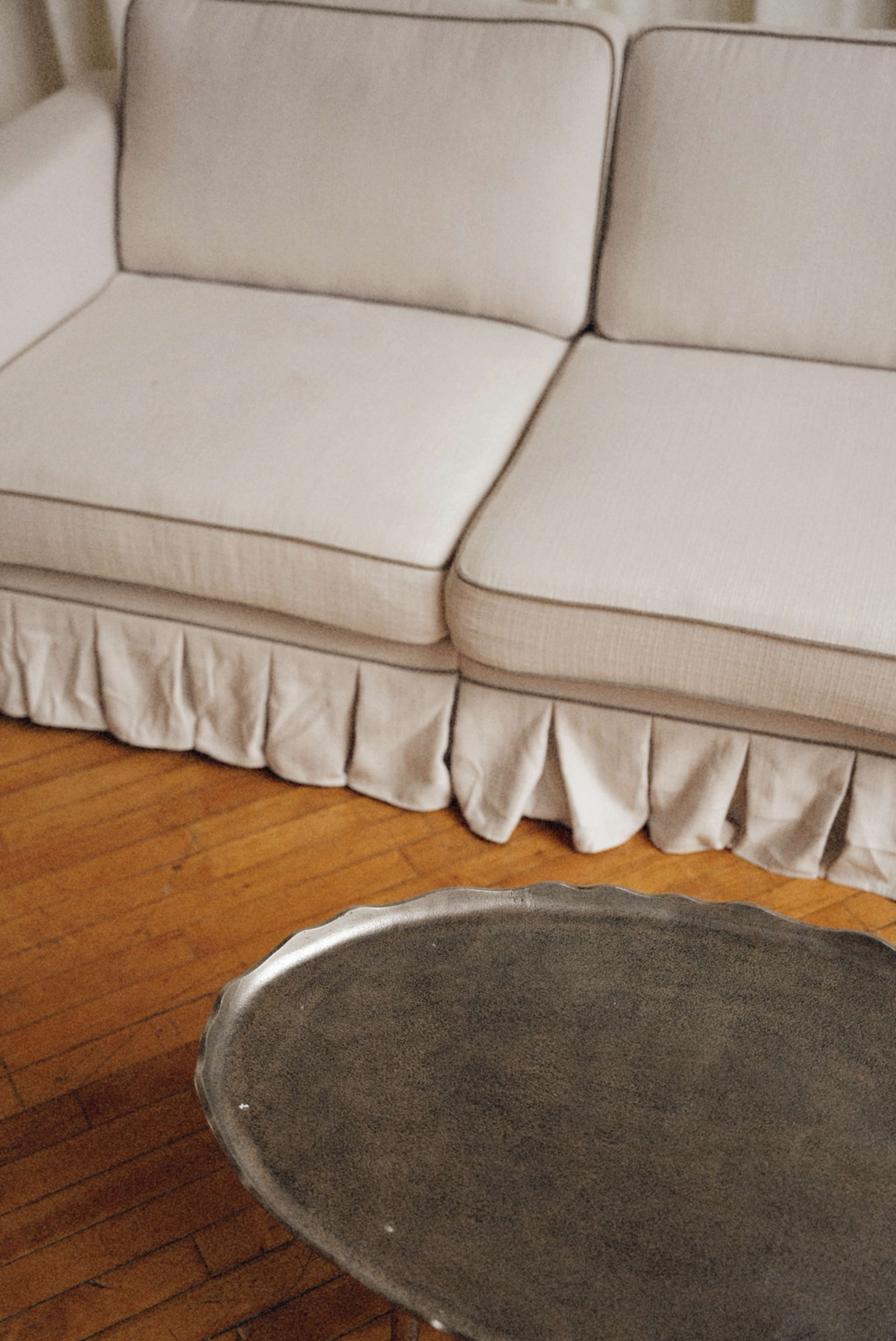 The image shows a light-colored couch with a pleated skirt positioned next to a round metallic side table on a wooden floor.