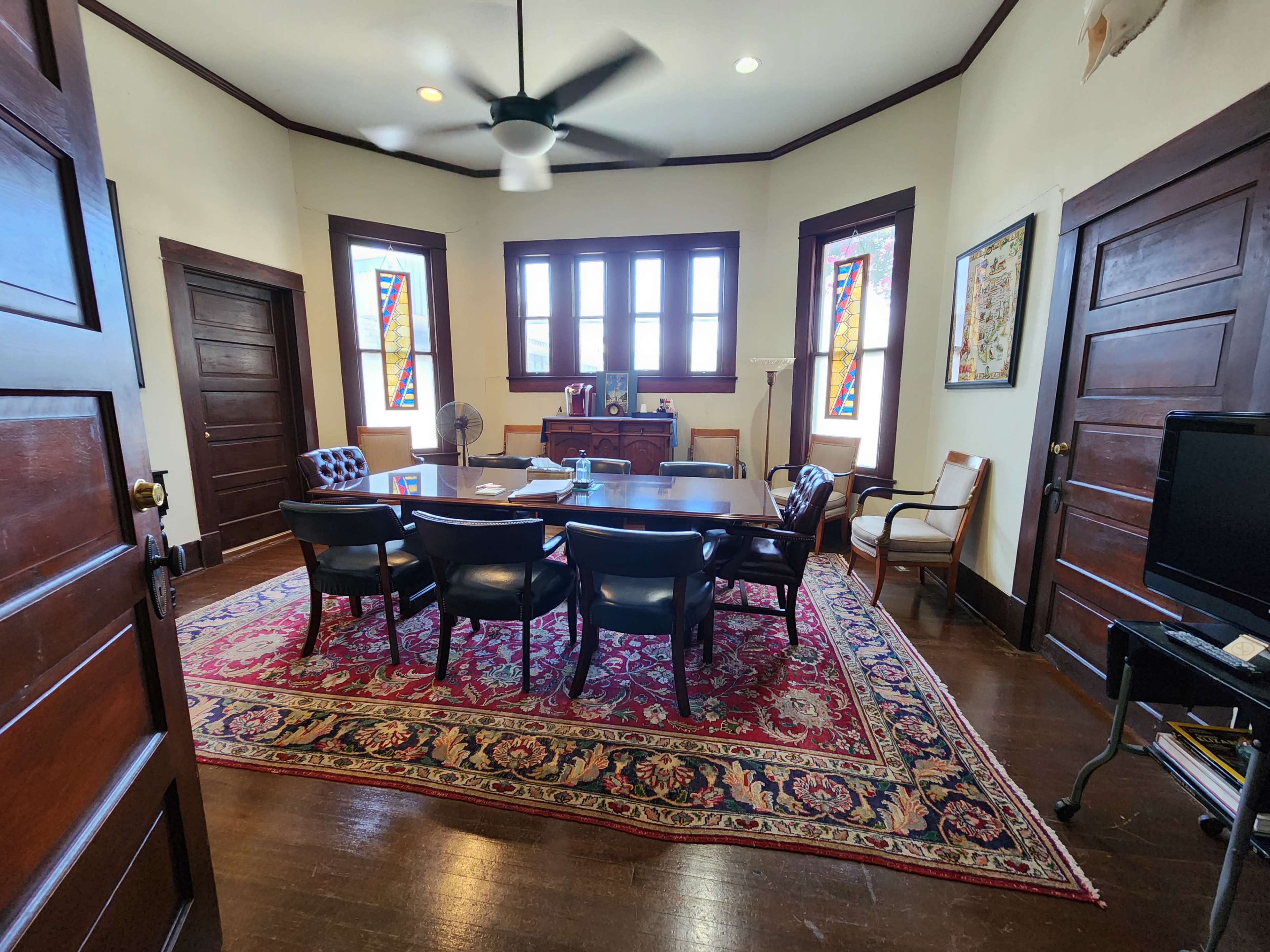 A conference room with a large table surrounded by black chairs, featuring stained glass windows and wooden doors.