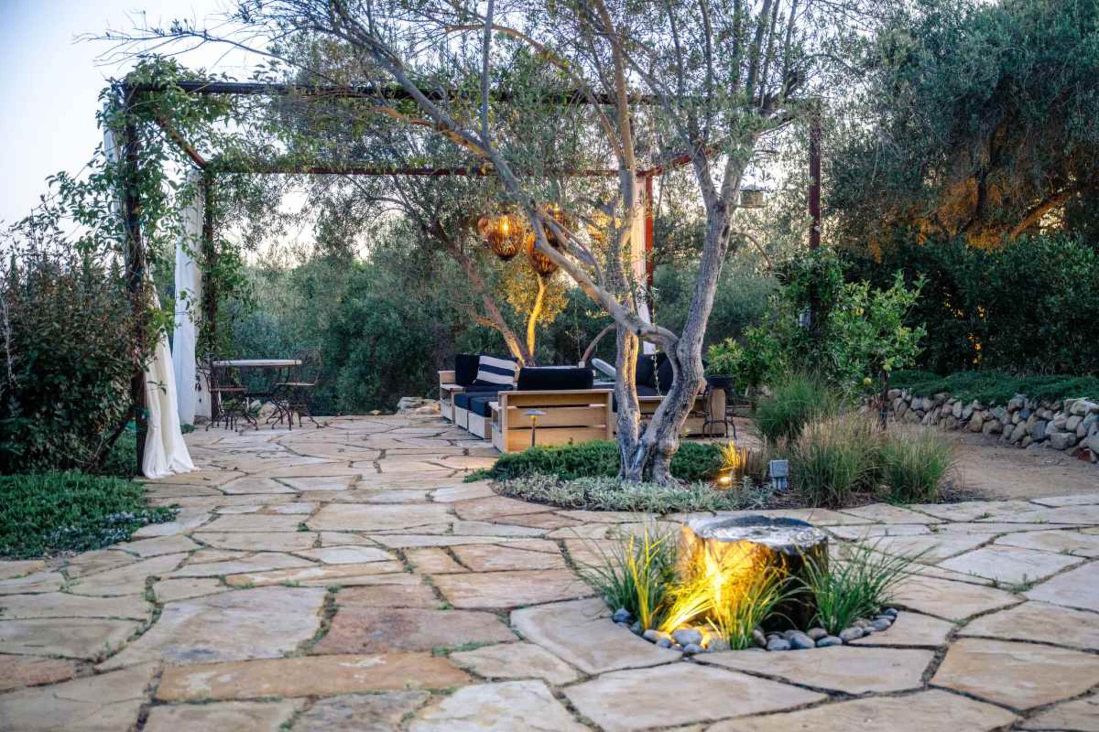 The image depicts a landscaped patio area with stone pathways, comfortable seating, and plants illuminated by soft lighting.