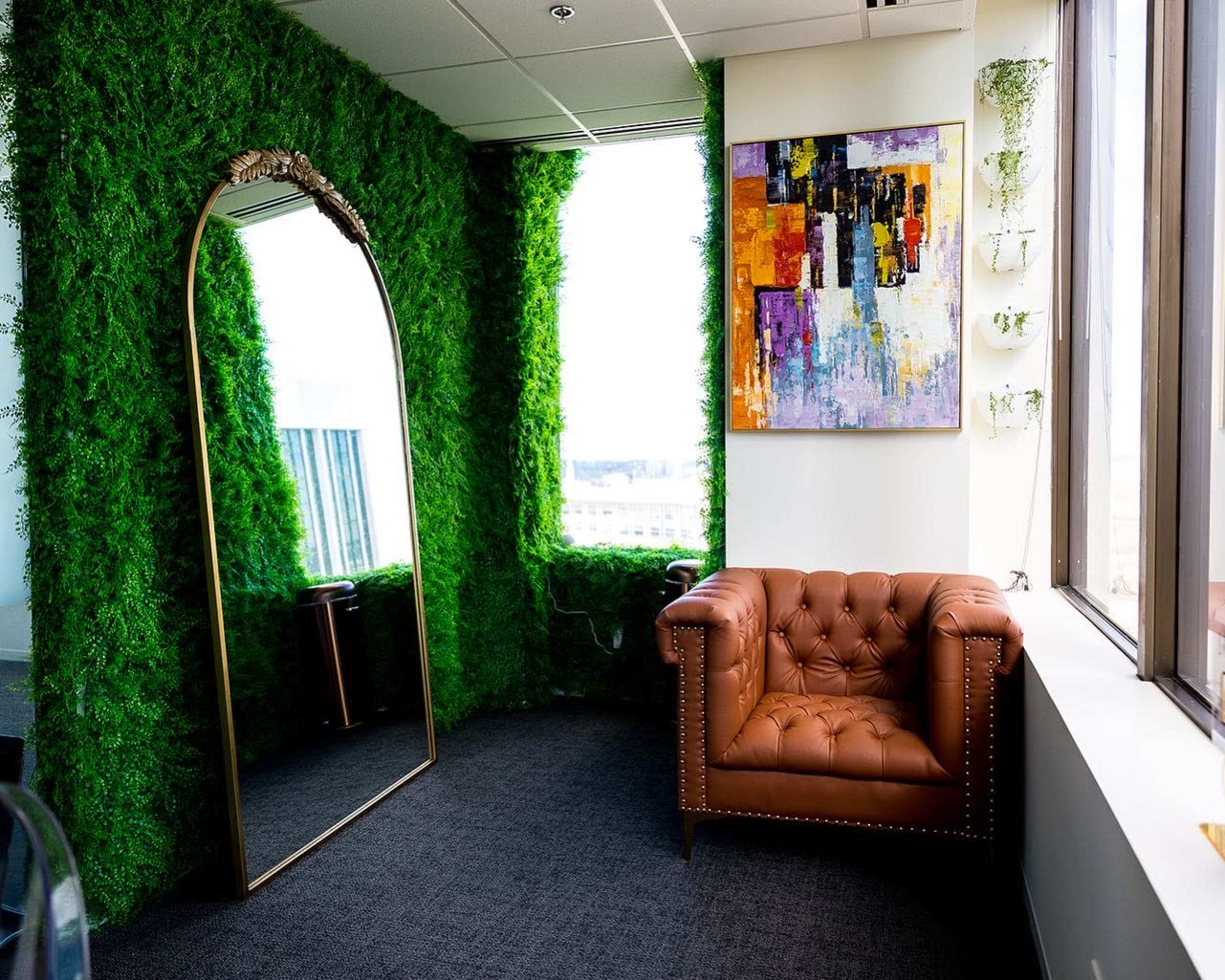 A corner of an office featuring a green wall of artificial grass, a large mirror, a colorful abstract painting, and a brown tufted leather armchair.