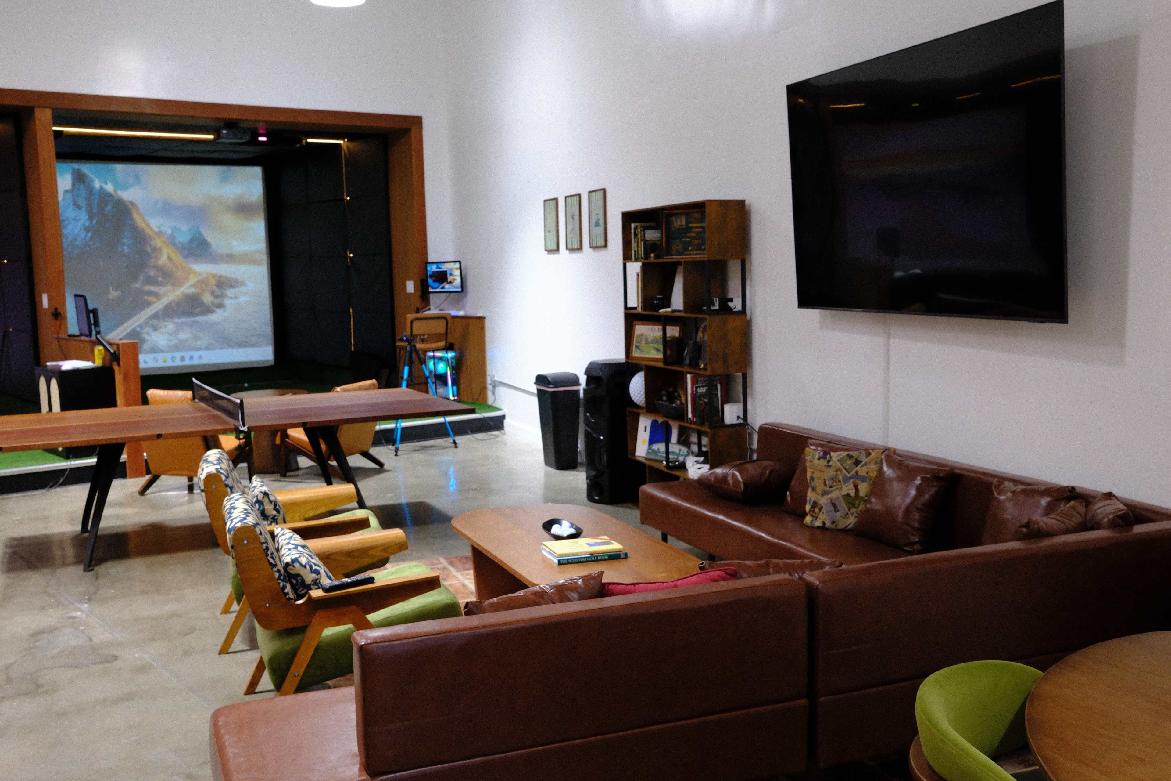 The image shows a modern lounge area with a brown leather couch, colorful chairs, a large wall-mounted TV, and a projector screen displaying a scenic landscape in a well-lit room.
