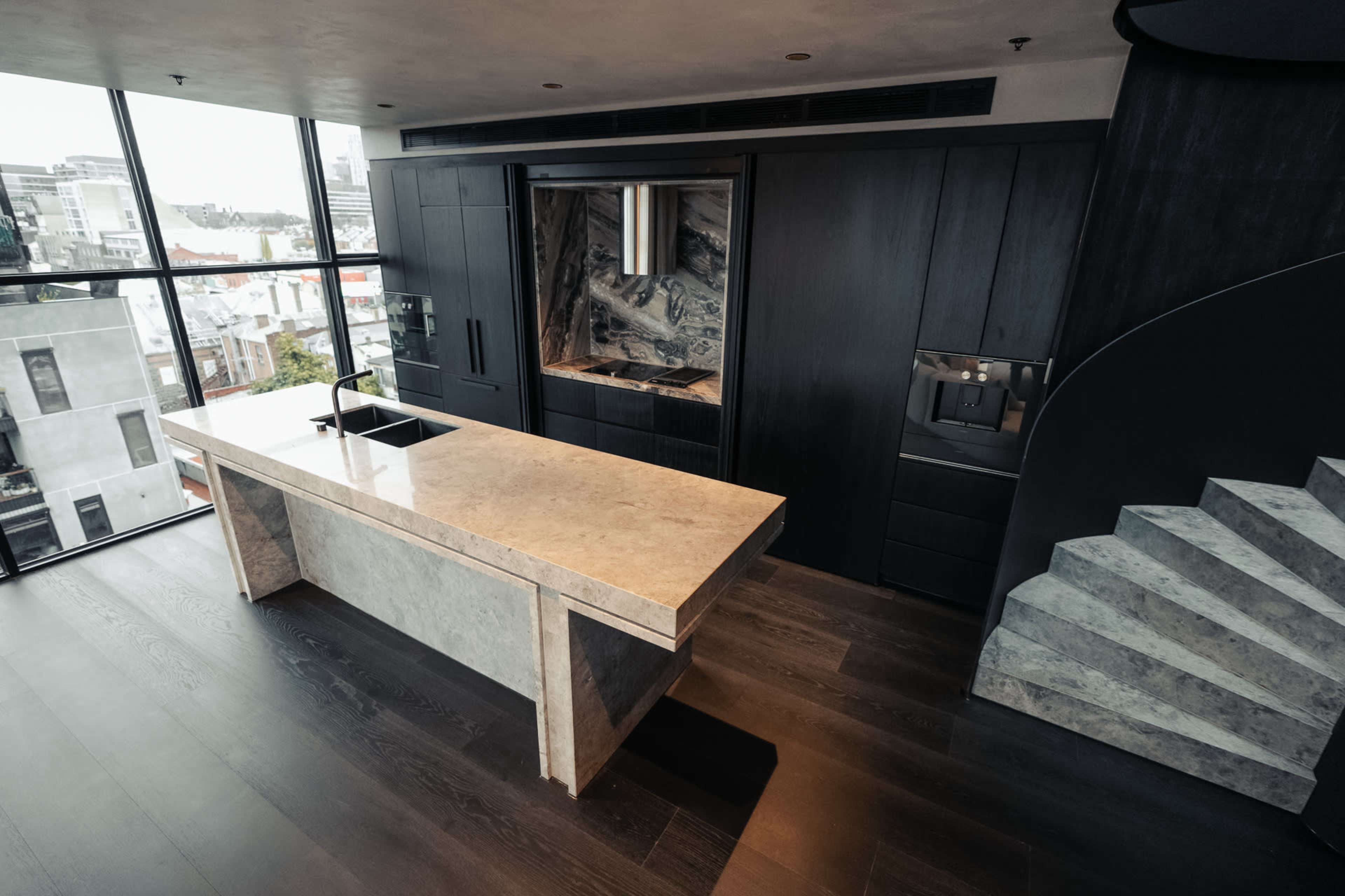 The image shows a modern kitchen featuring a large stone island, dark cabinetry, and a curved staircase in the background.