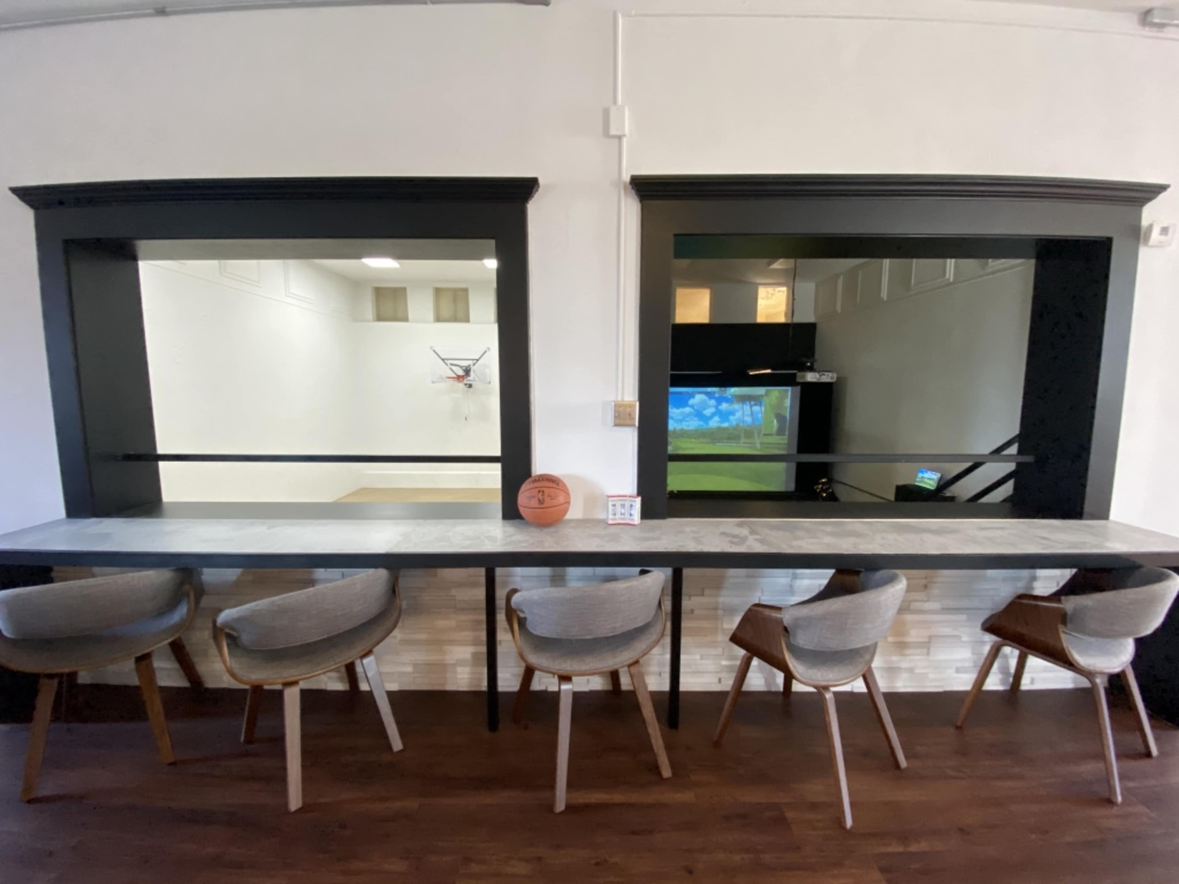 A seating area with five chairs arranged at a bar counter, leading to two large windows that overlook a basketball court and a projector screen.