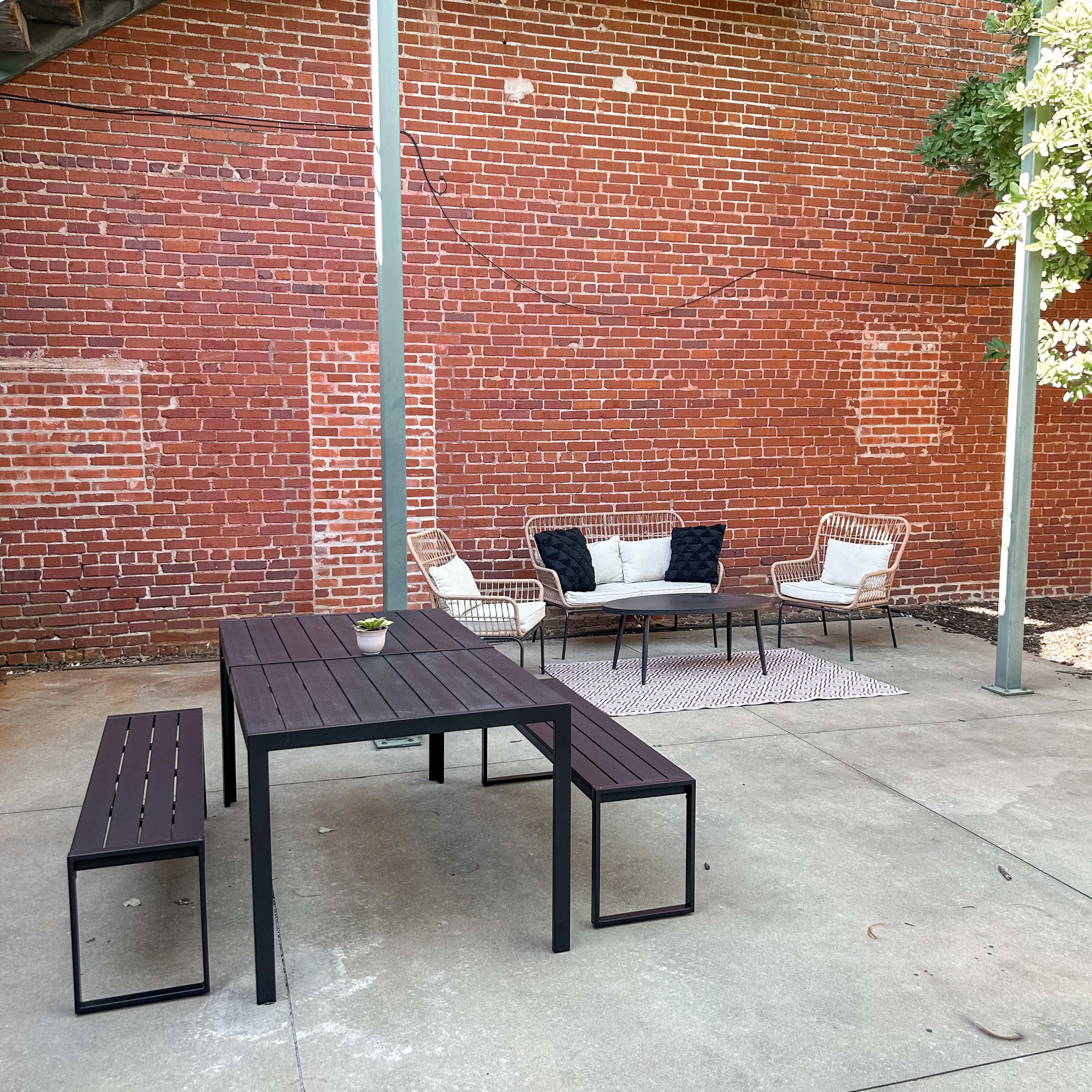 A metal table with two benches and a seating area of chairs is set on a concrete patio against a brick wall.