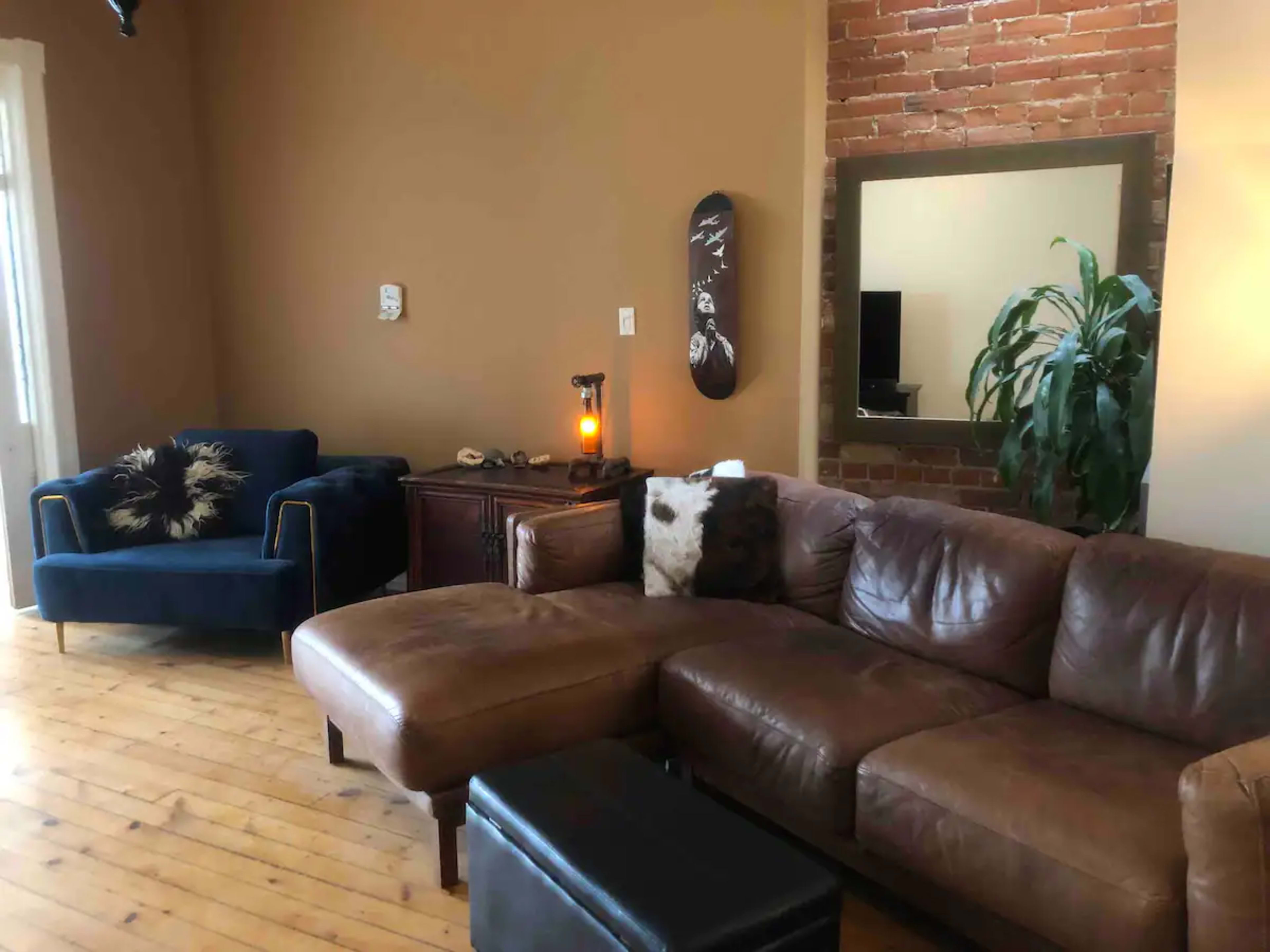 The image shows a cozy living room with a sectional leather couch, a blue armchair, a wooden cabinet, and a large mirror on the wall, complemented by a decorative plant and exposed brick.