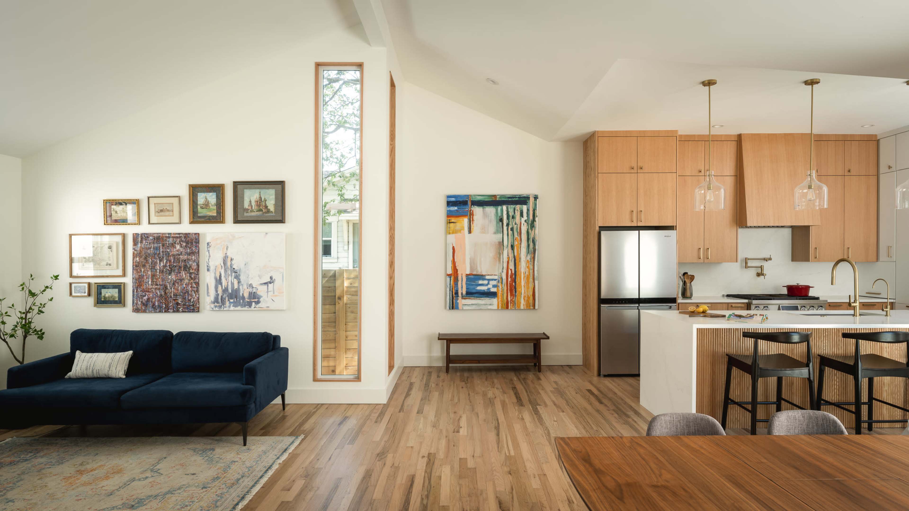The image shows a modern living space featuring a dark blue sofa, framed artwork on the walls, a wooden dining table, and a kitchen with stainless steel appliances.