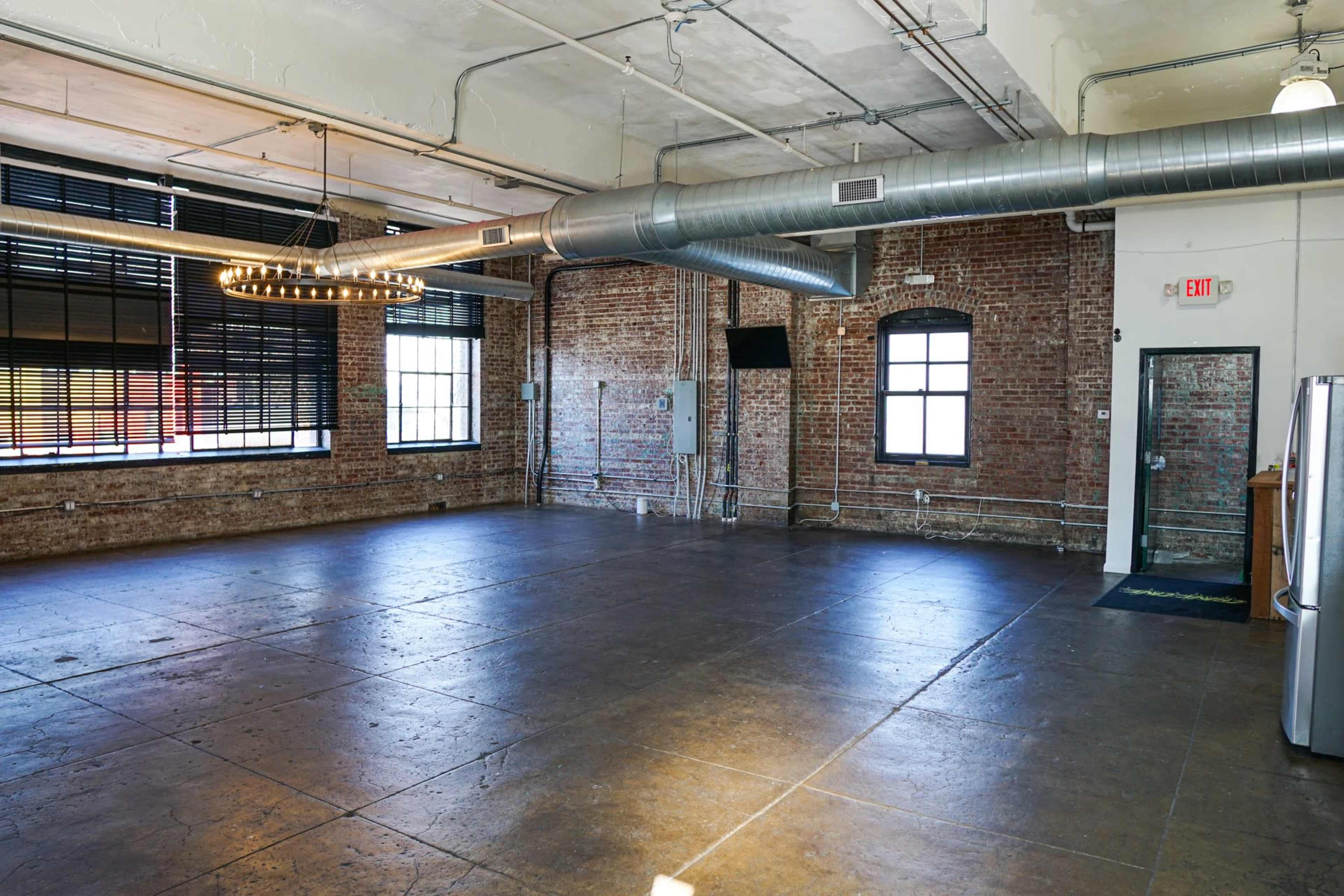 An empty industrial-style room with exposed brick walls, large windows, and a metal duct running along the ceiling.