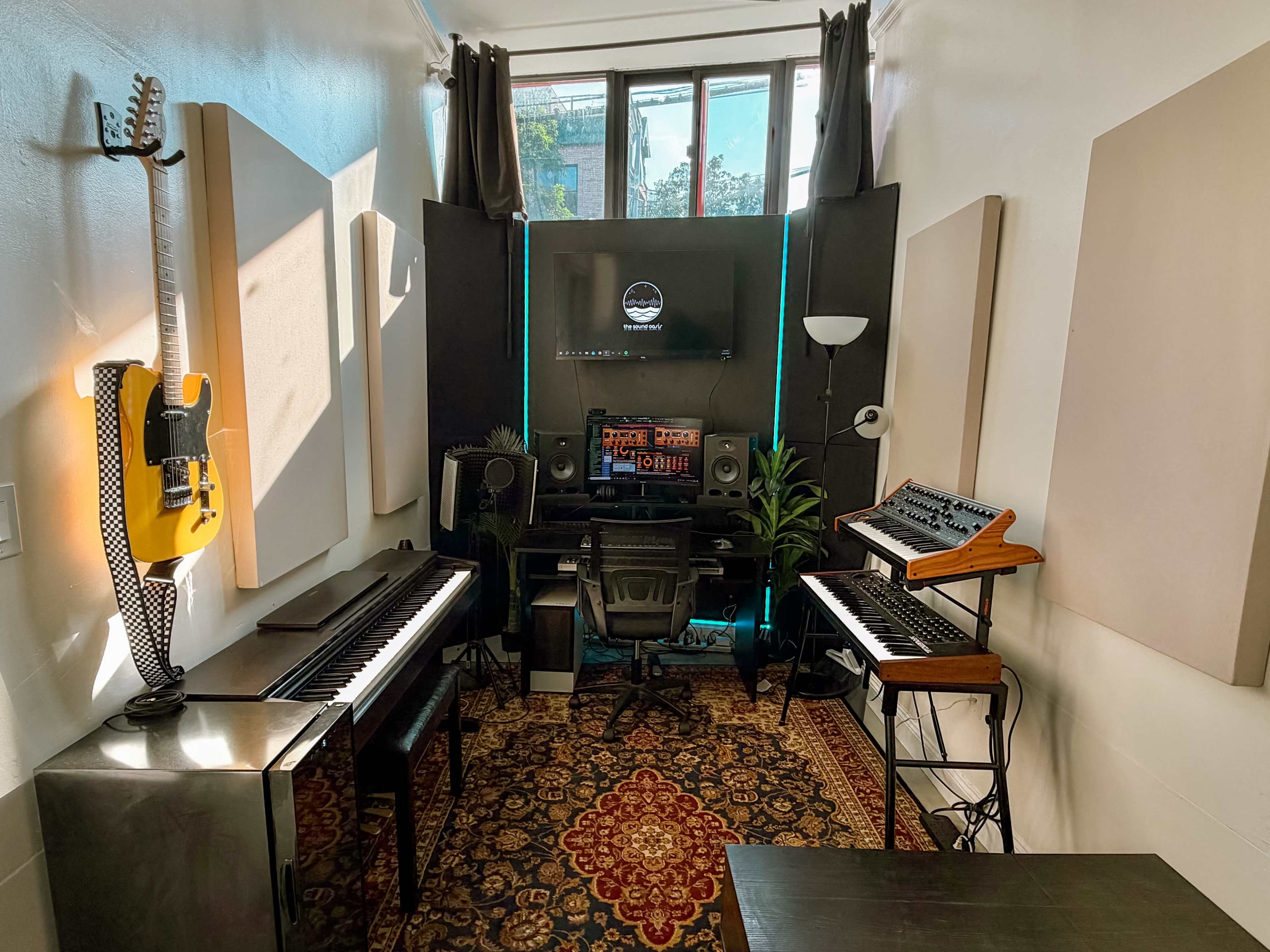 The image shows a small music production studio featuring two keyboards, a computer setup with a monitor, a synthesizer, and soundproofing panels on the walls.