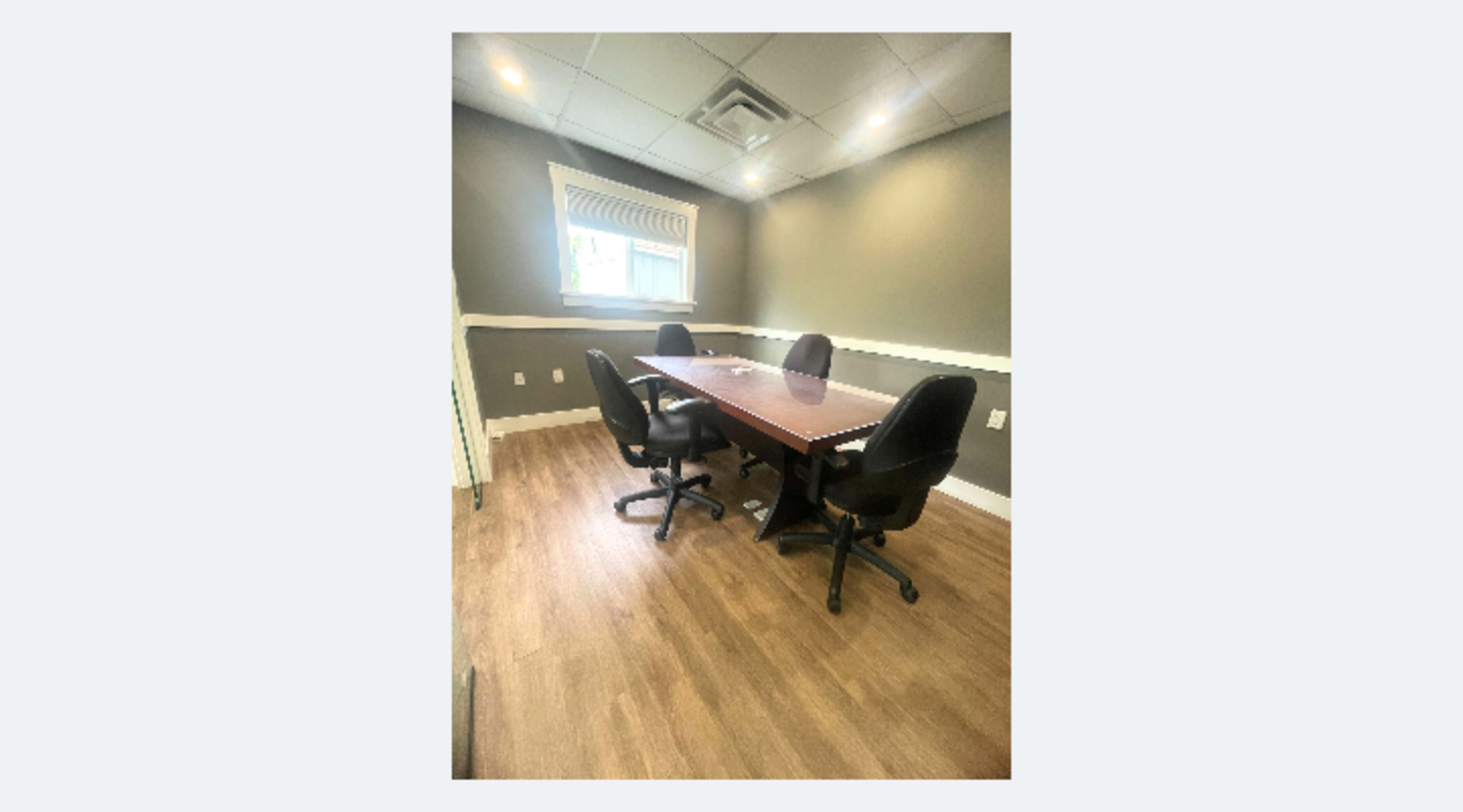 The image shows a small conference room with a wooden table and four black chairs, illuminated by natural light from a window.