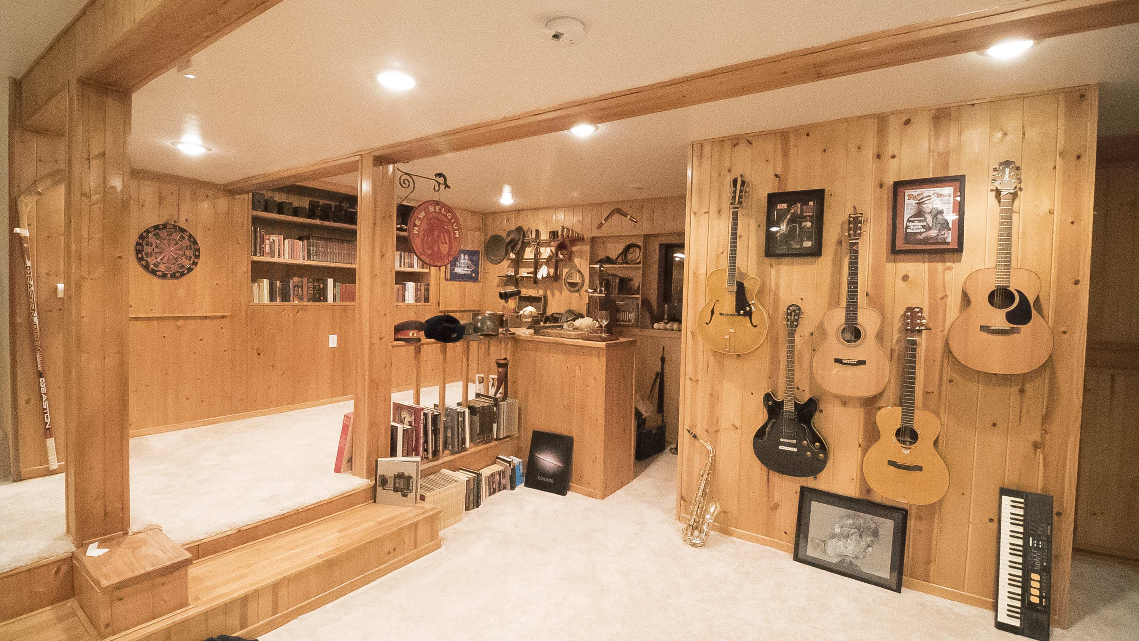 The image shows a wooden-paneled basement with a small kitchen area, shelves filled with books and decorative items, and various musical instruments hanging on the wall.