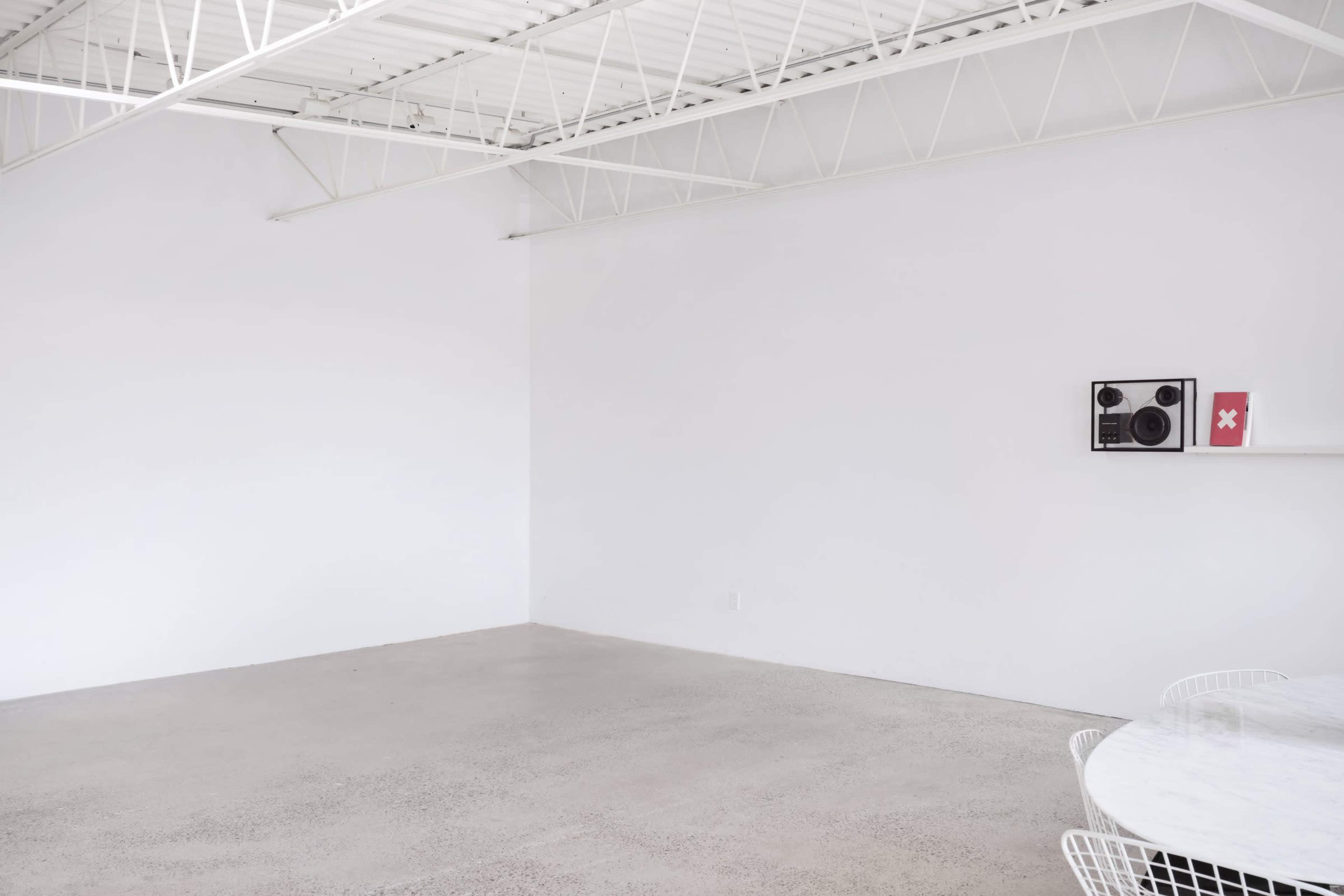 The image shows a minimalist interior space with white walls, a concrete floor, and a small shelf displaying a sound system and a red book.