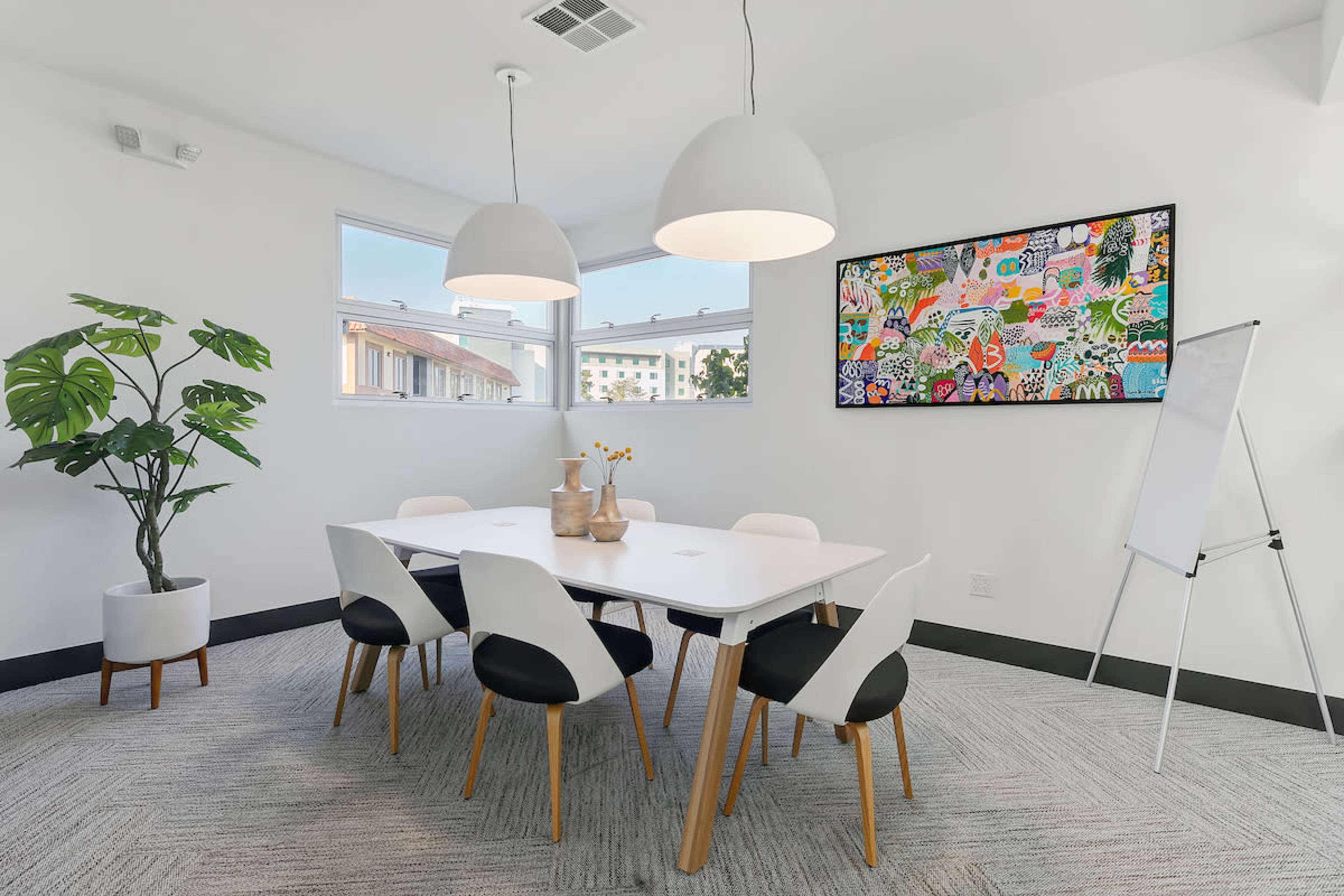 A brightly lit meeting room features a large white table surrounded by six chairs, with a plant and colorful artwork on the walls.