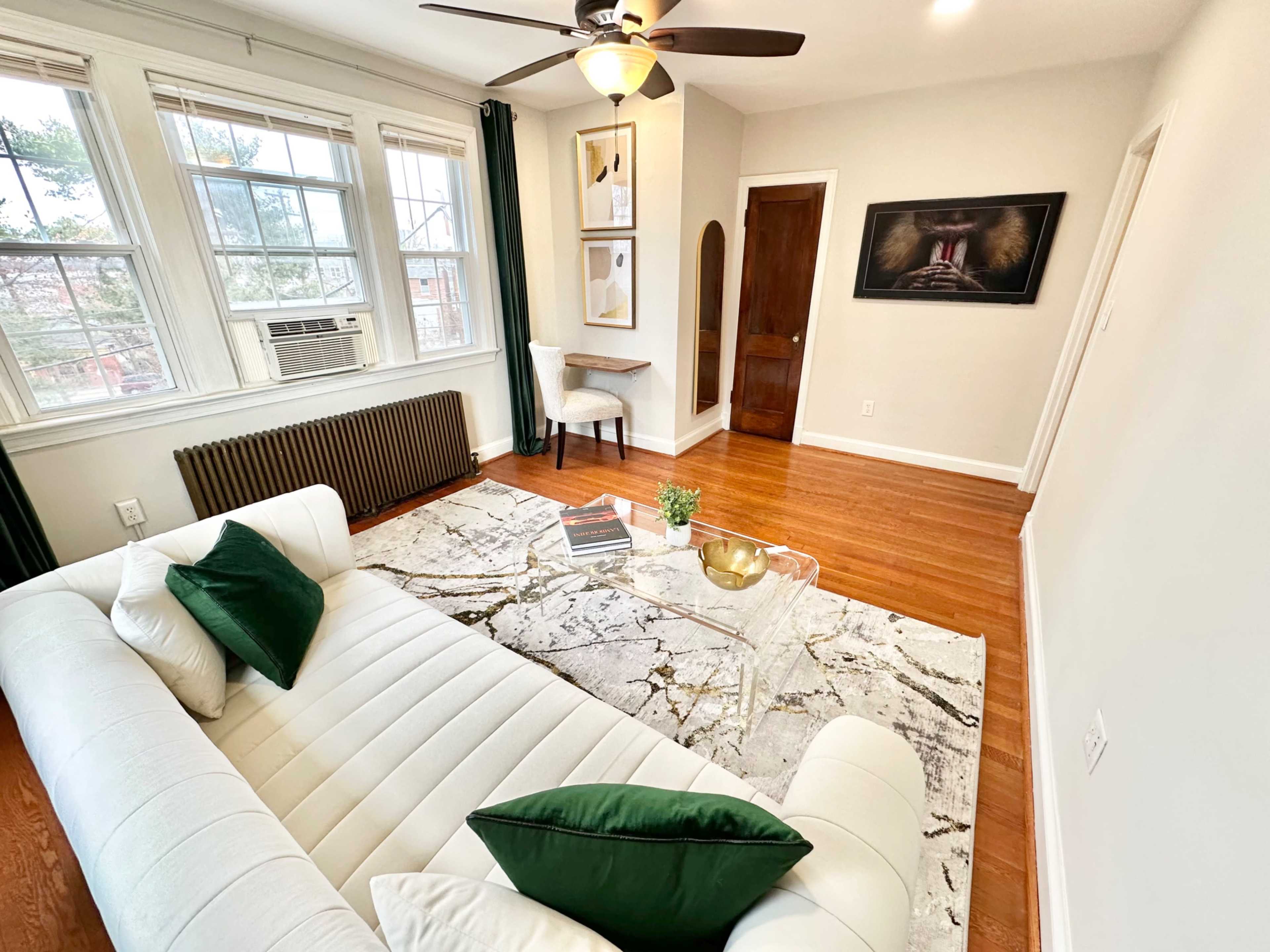 A bright living room features a white sofa with green pillows, a patterned rug, and a small table, all complemented by large windows and hardwood floors.