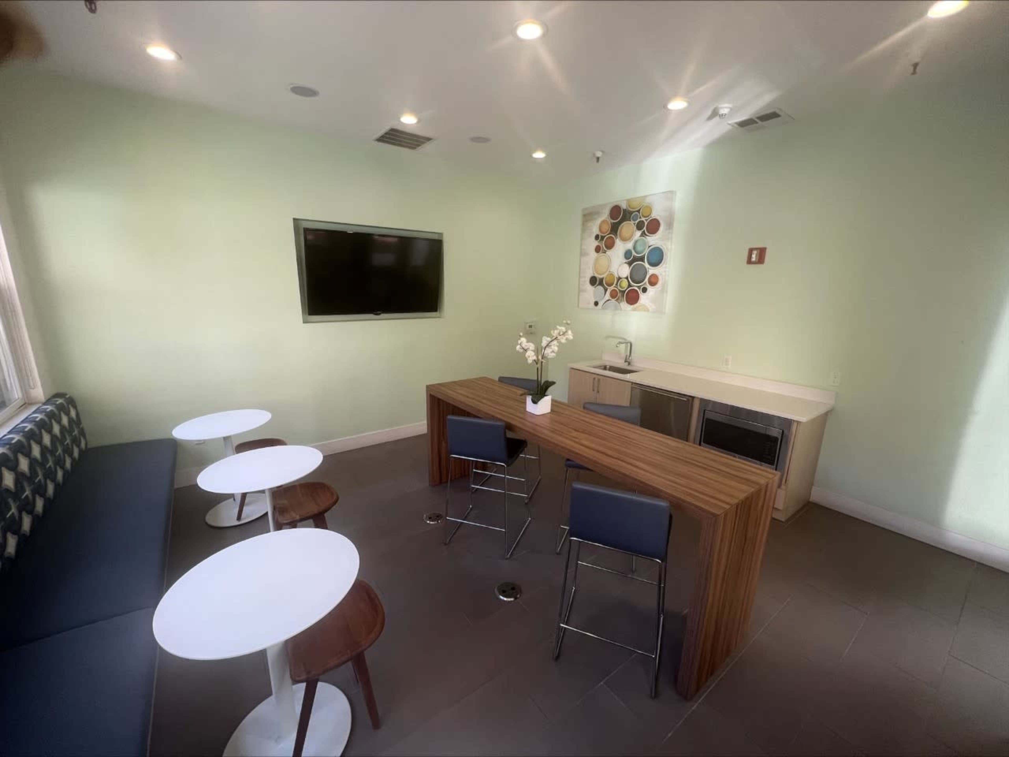 A modern lounge area with a kitchenette, featuring a wooden table and four seating areas consisting of two small round tables and two chairs.