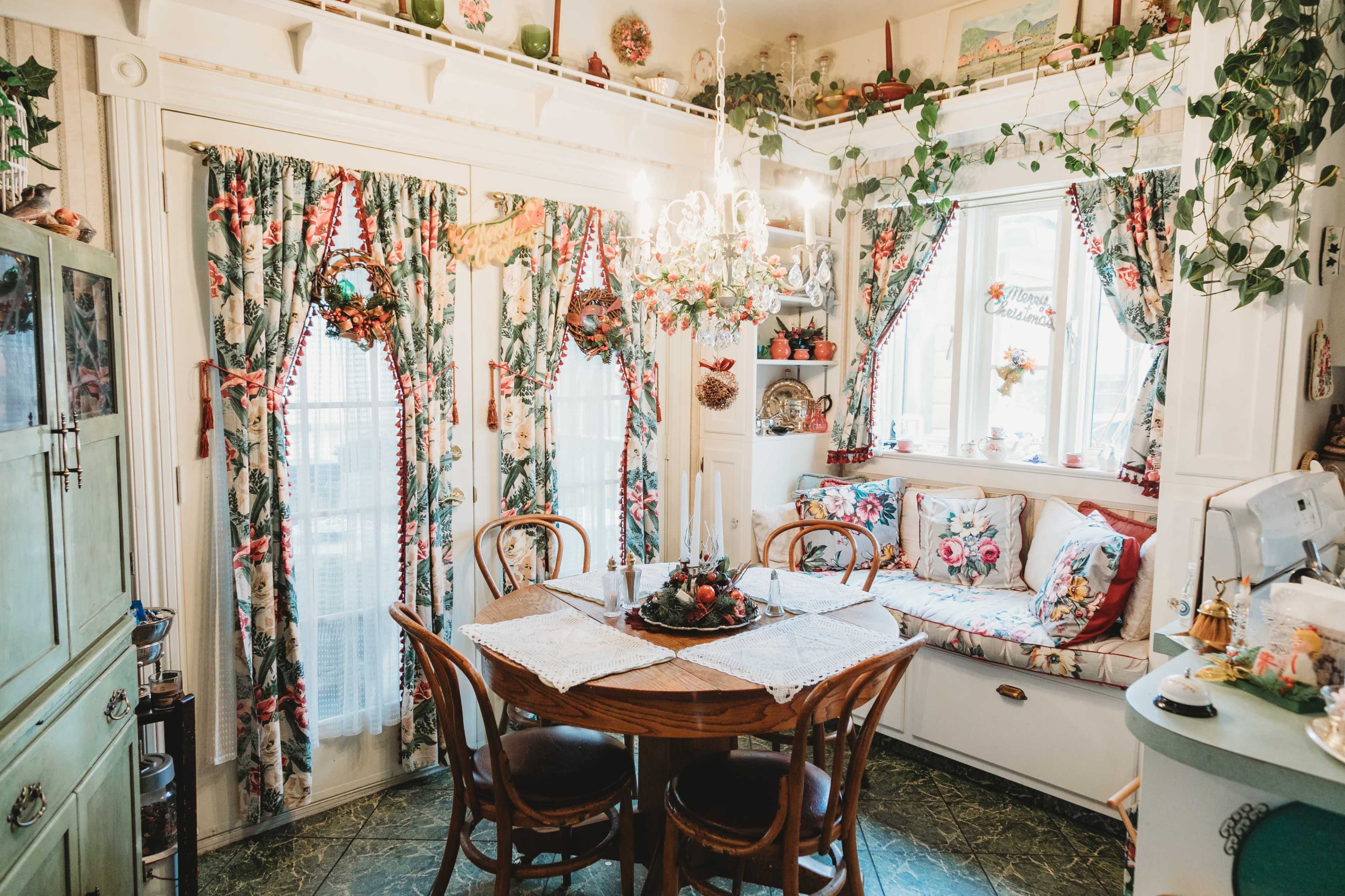 A cozy kitchen features a wooden dining table surrounded by chairs, with floral curtains and decorative plants adding a touch of nature to the space.