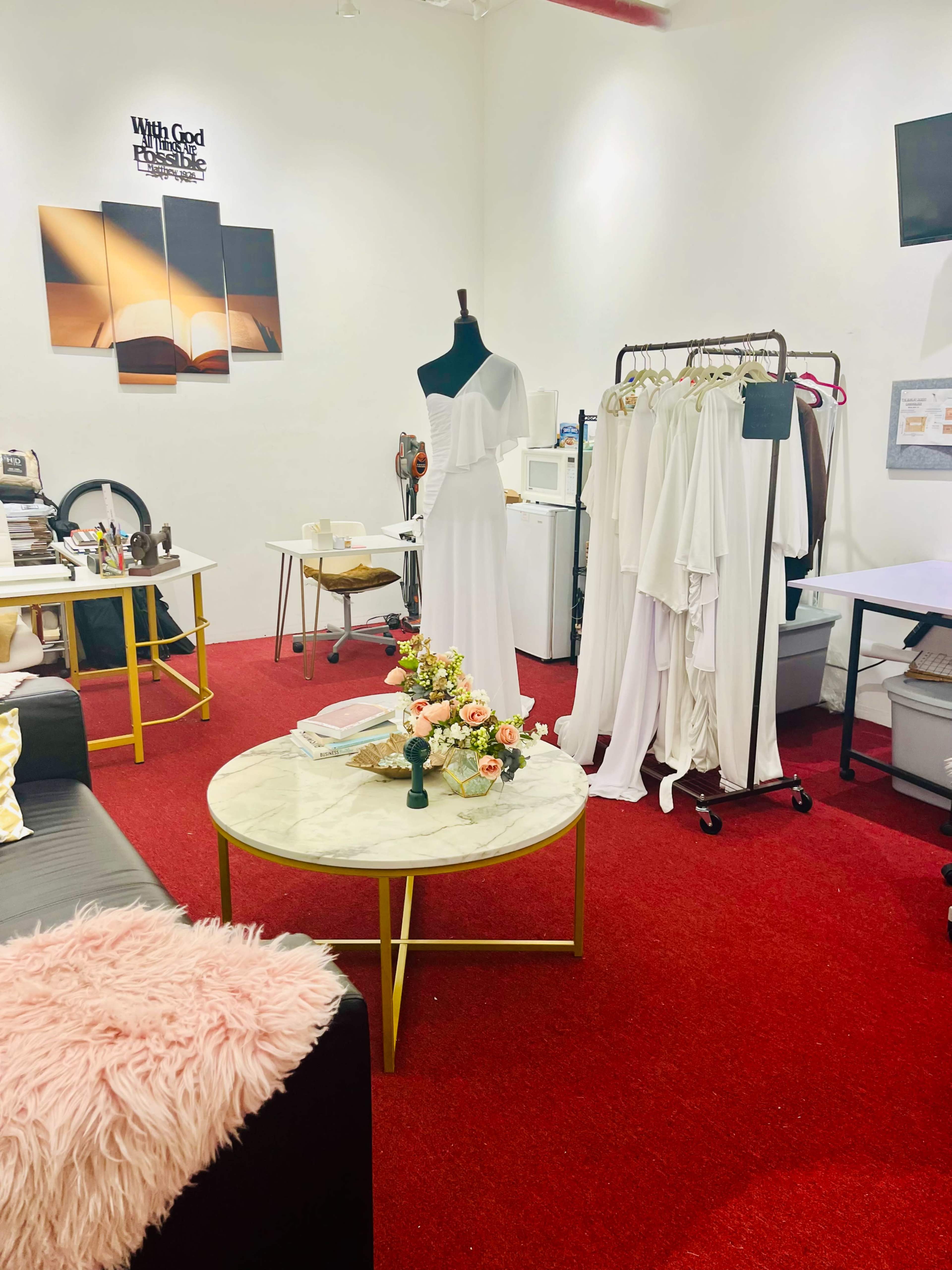 The room features a red carpeted floor with a table at the center surrounded by clothing racks and a mannequin displaying a white dress.