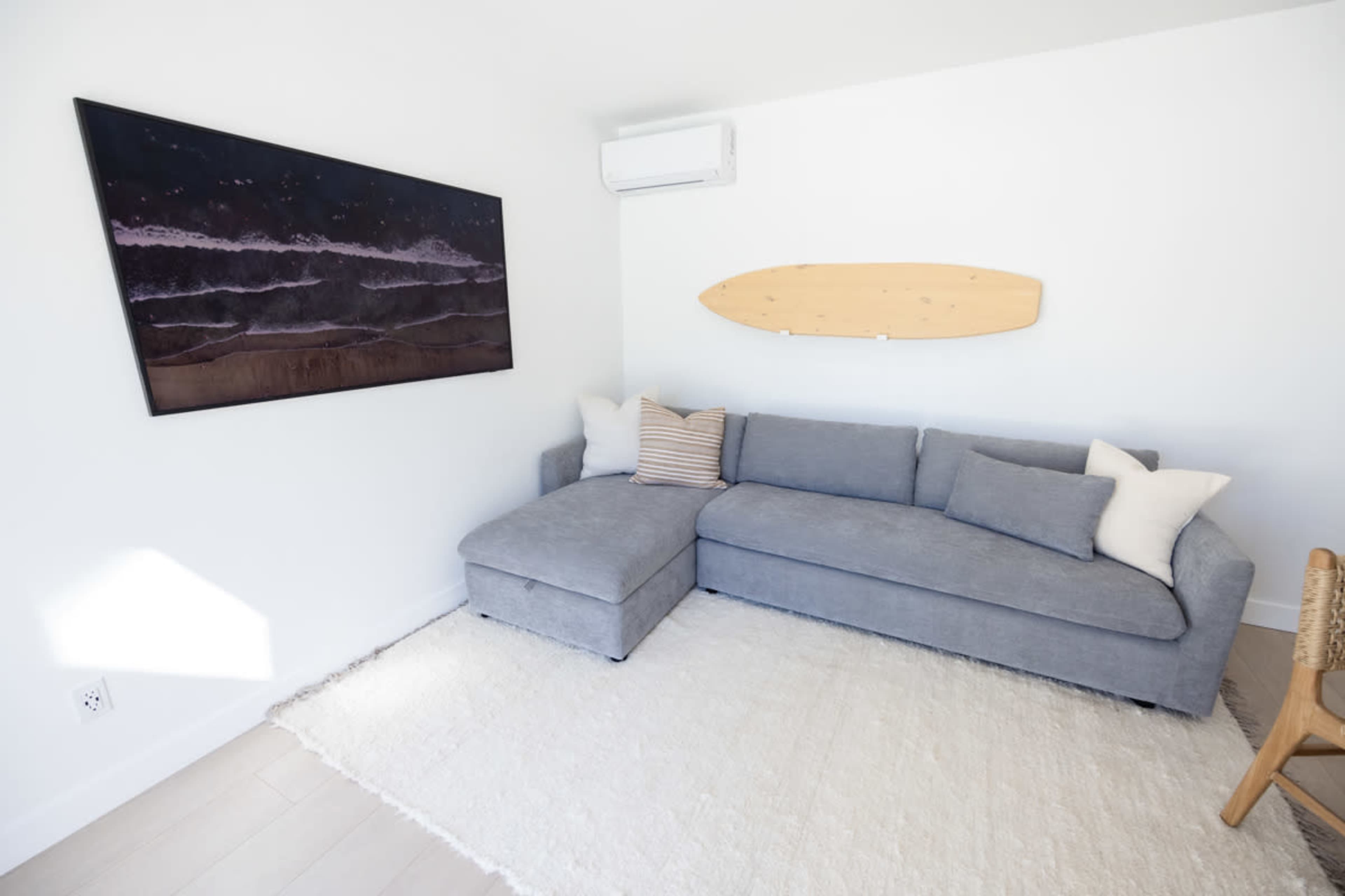 A light-filled living room features a gray sectional sofa with pillows, a large artwork on the wall, and a wooden surfboard mounted above.