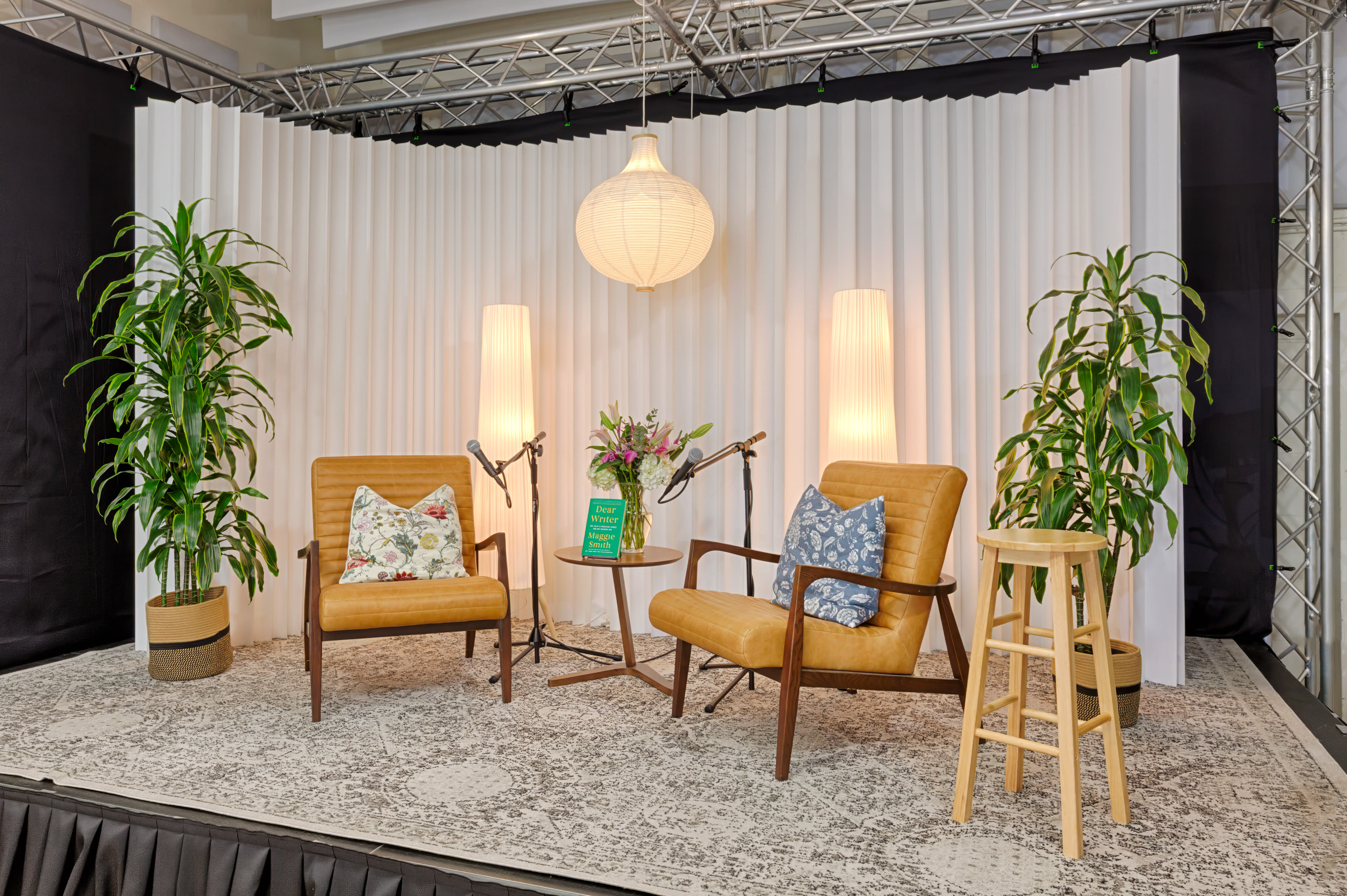 The image shows an arranged stage setup with two yellow armchairs, a wooden stool, plants, and decorative lighting against a white backdrop.