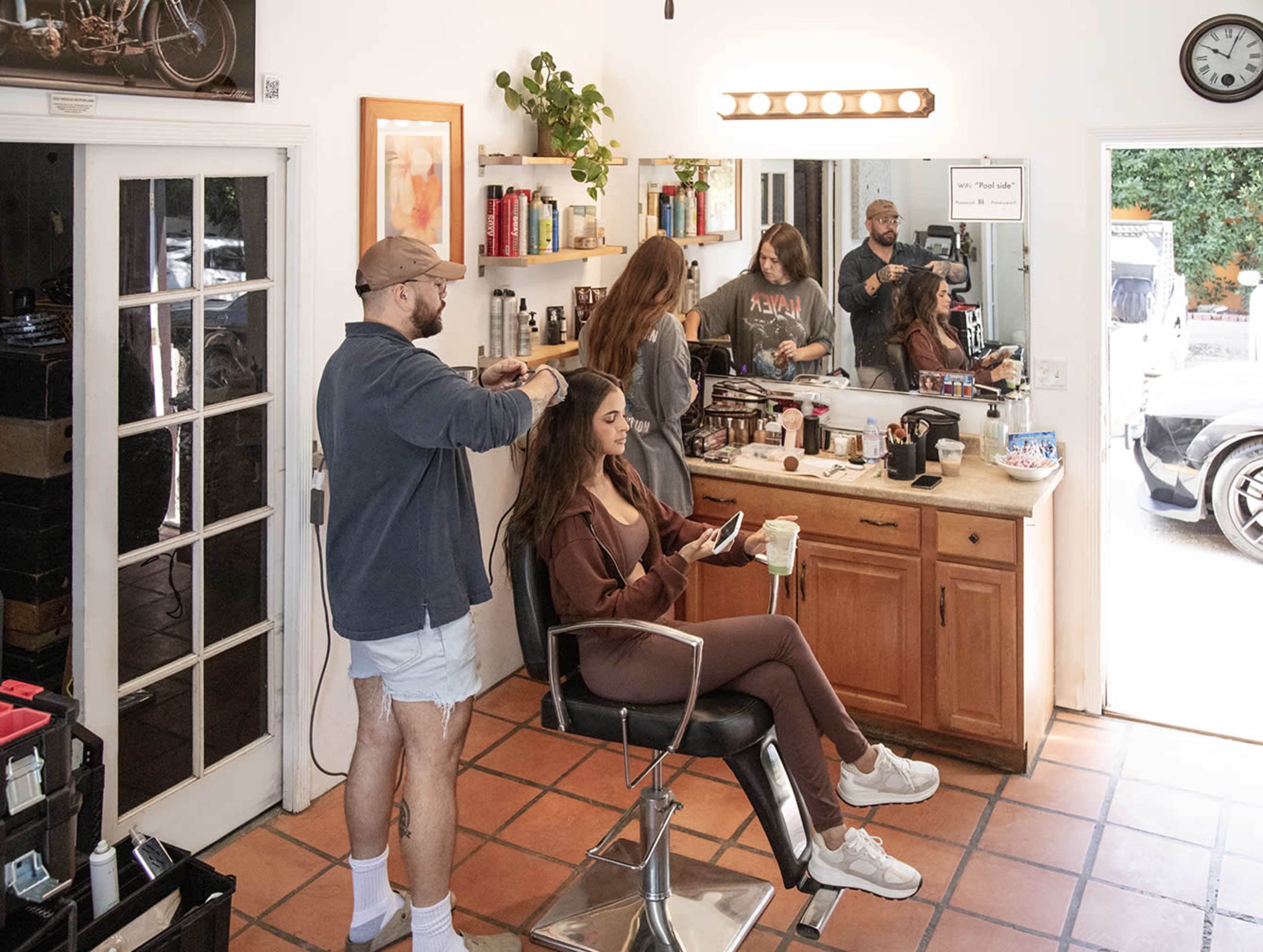 A hairstylist works on a woman's hair in a salon, while another woman sits nearby looking at her phone and a barber is visible in the mirror.