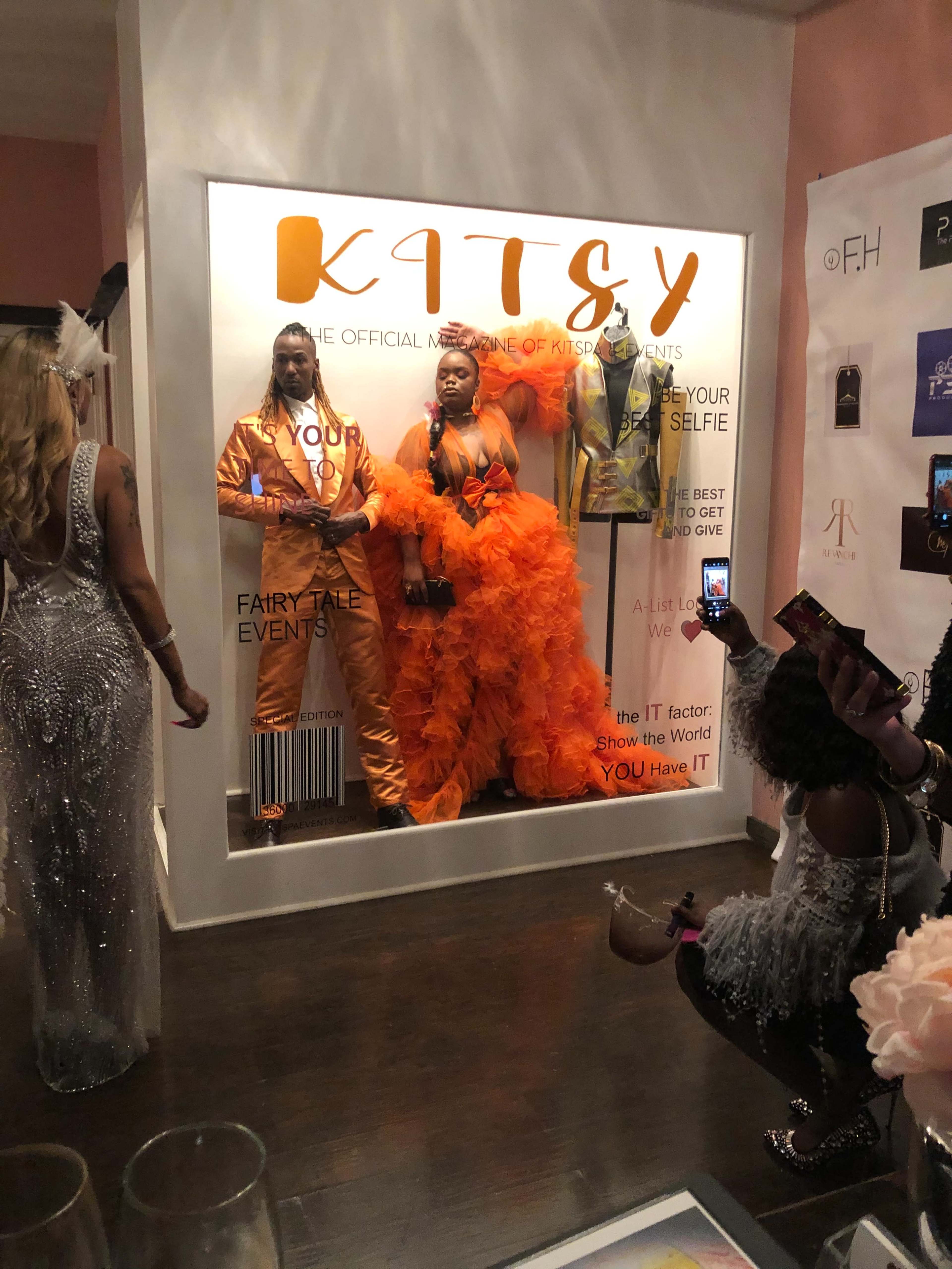 A glass display showcases three individuals dressed in vibrant orange outfits, posing in front of a backdrop featuring the word "KITSY" and event promotional materials.