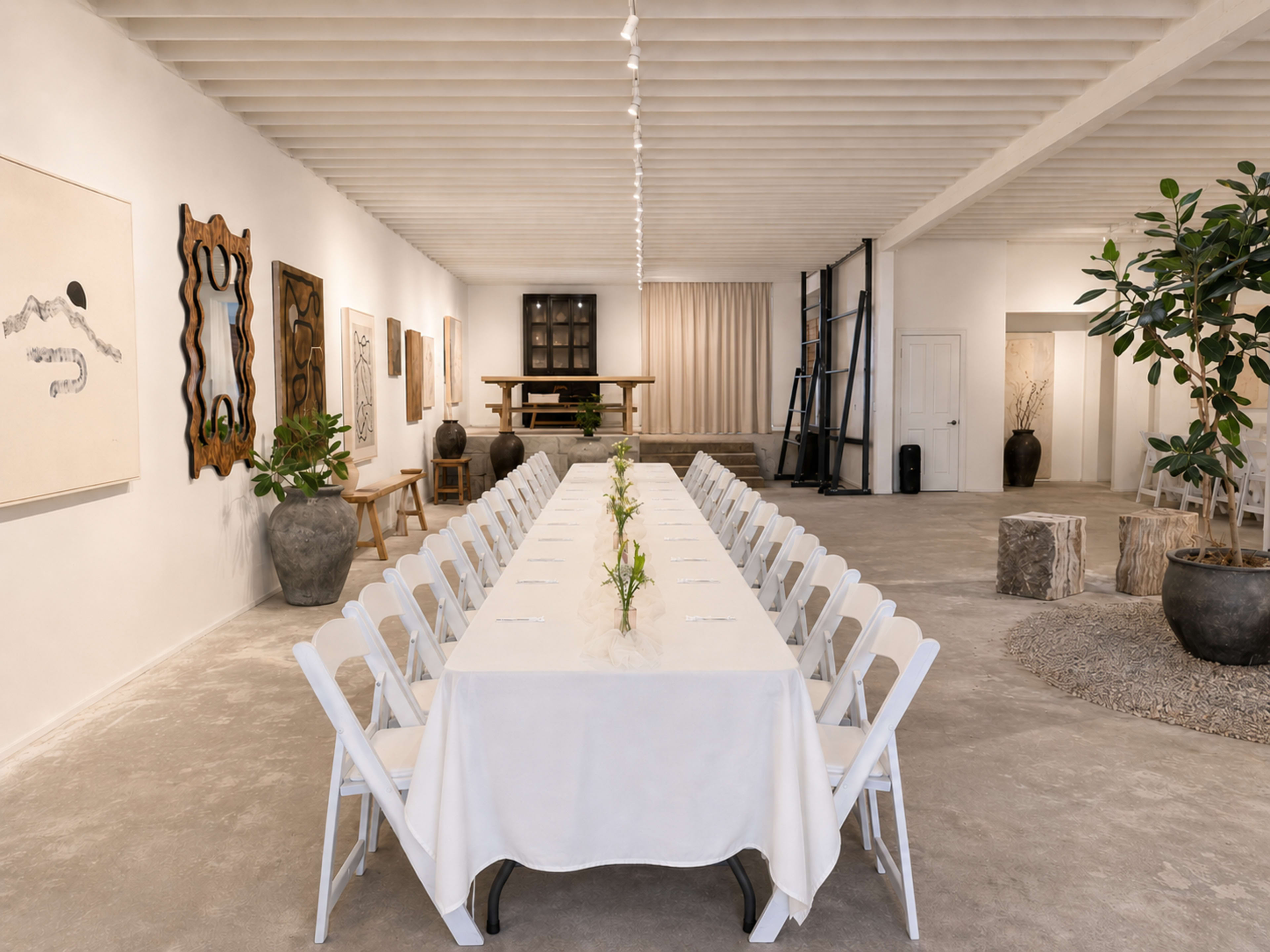 A long table with white tablecloths is set up in a spacious, art-filled room with light-colored walls and a simple, minimalist decor.
