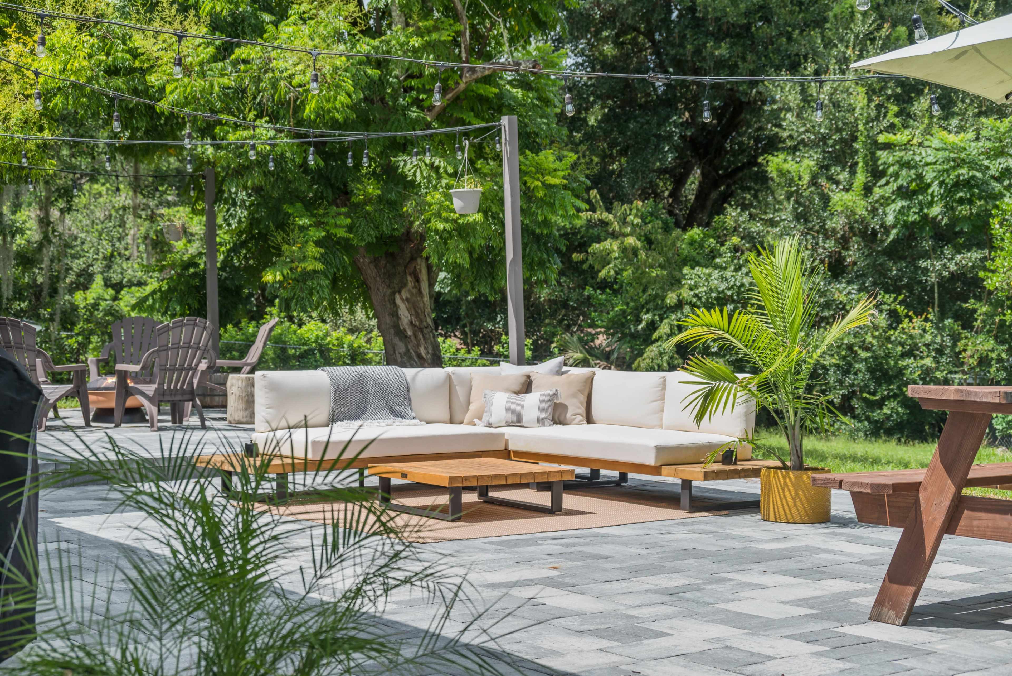 An outdoor seating area with a sectional sofa, wooden tables, and potted plants on a stone patio surrounded by greenery.