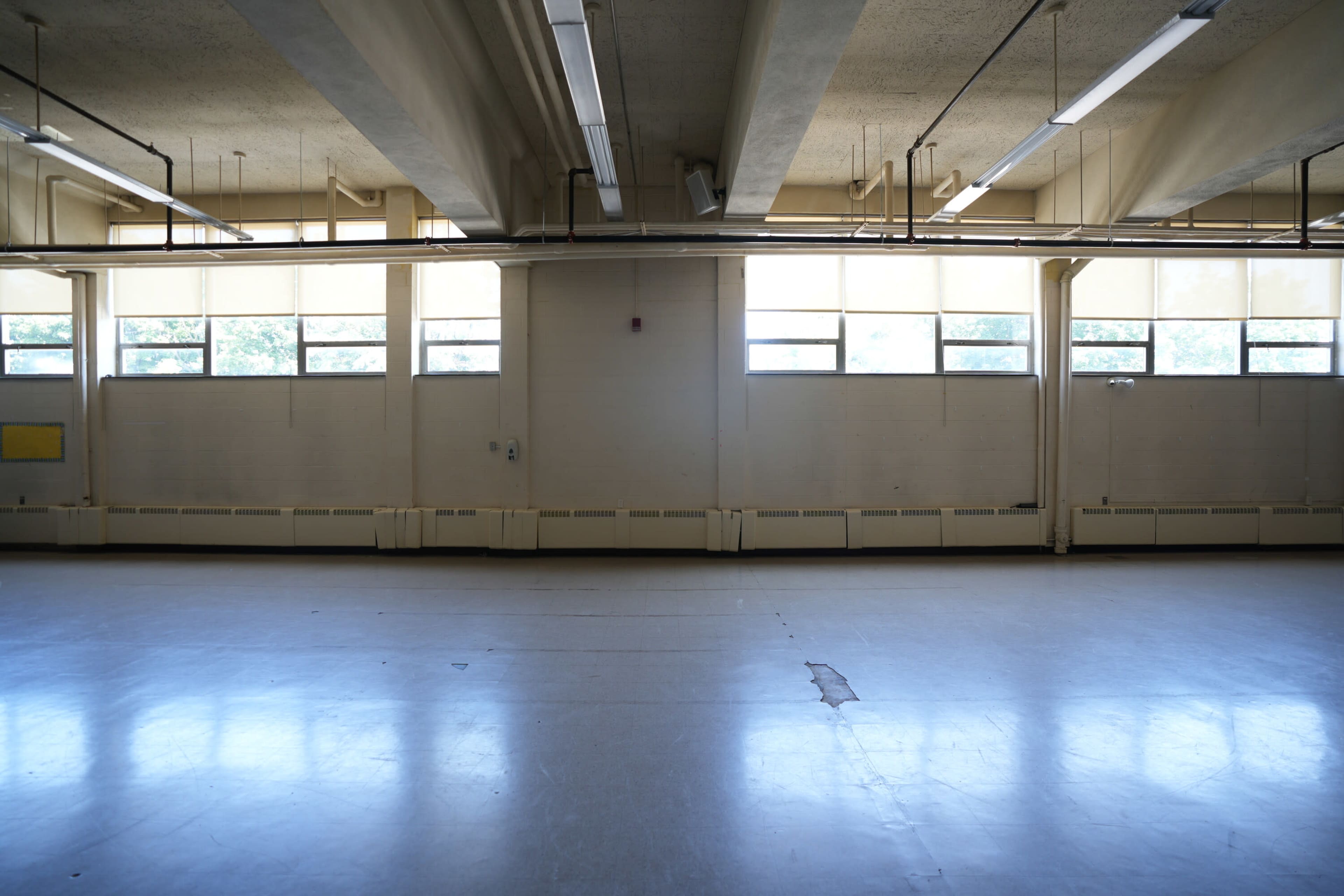 The image shows a spacious, empty room with large windows letting in natural light and a polished floor.