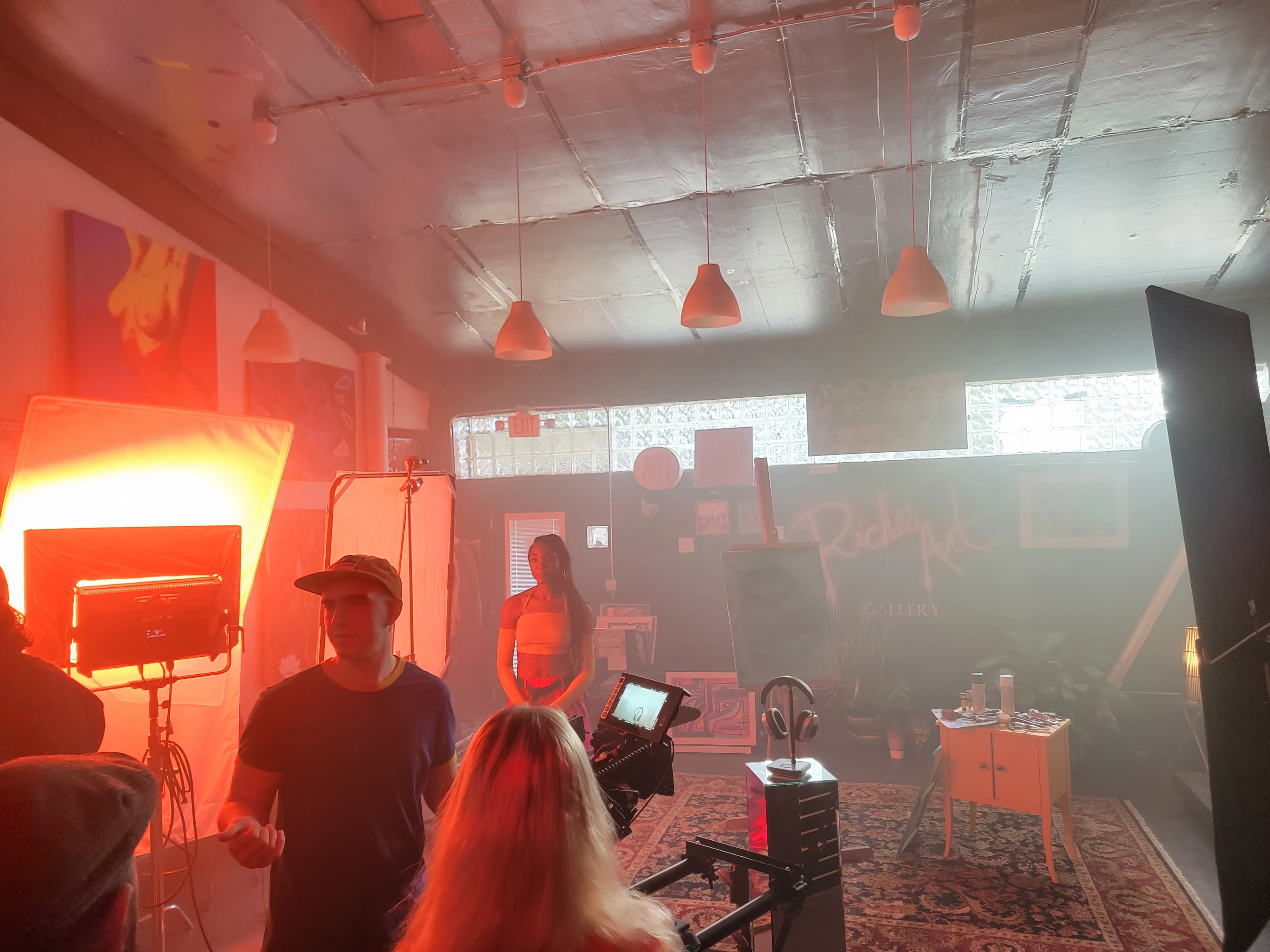 A well-lit gallery scene features a model posing in front of a backdrop, surrounded by production equipment and crew members.