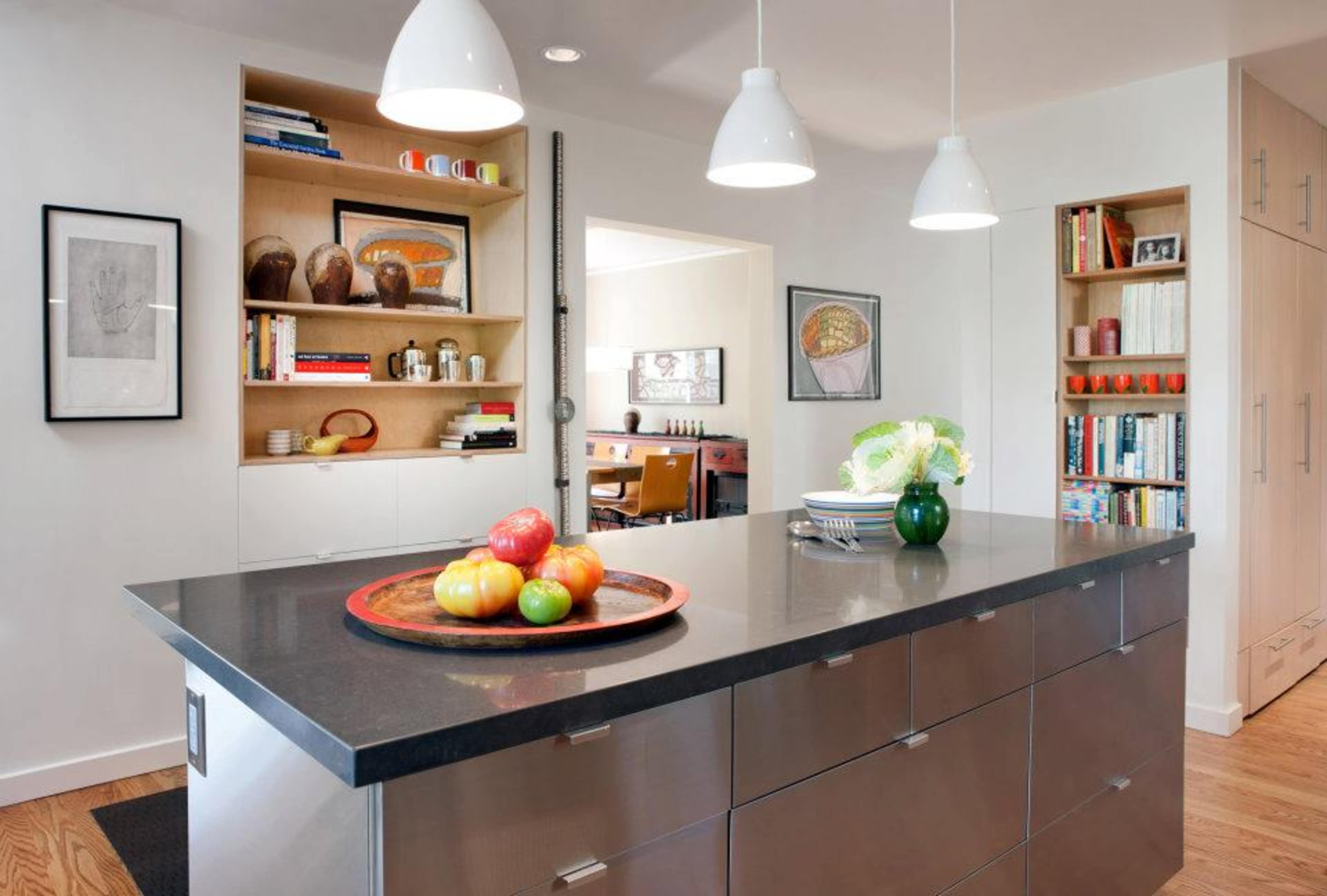 A modern kitchen featuring a central island with a bowl of colorful fruit, stainless steel cabinetry, and open shelving displaying various kitchen items.