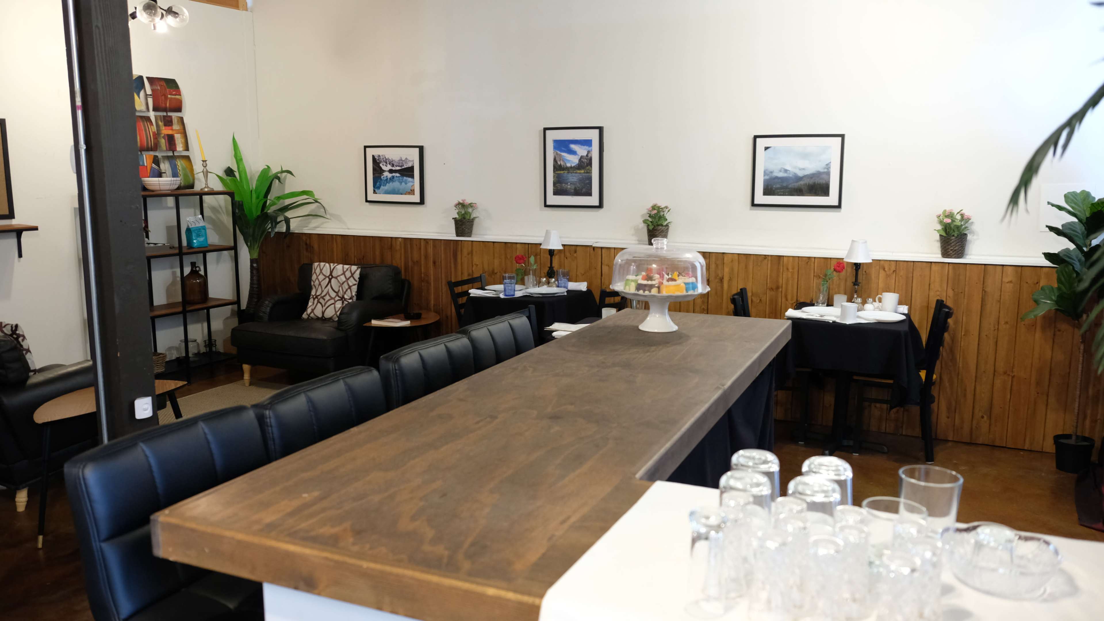 A cozy café interior with a long wooden table, black chairs, and framed landscape photographs on the walls.