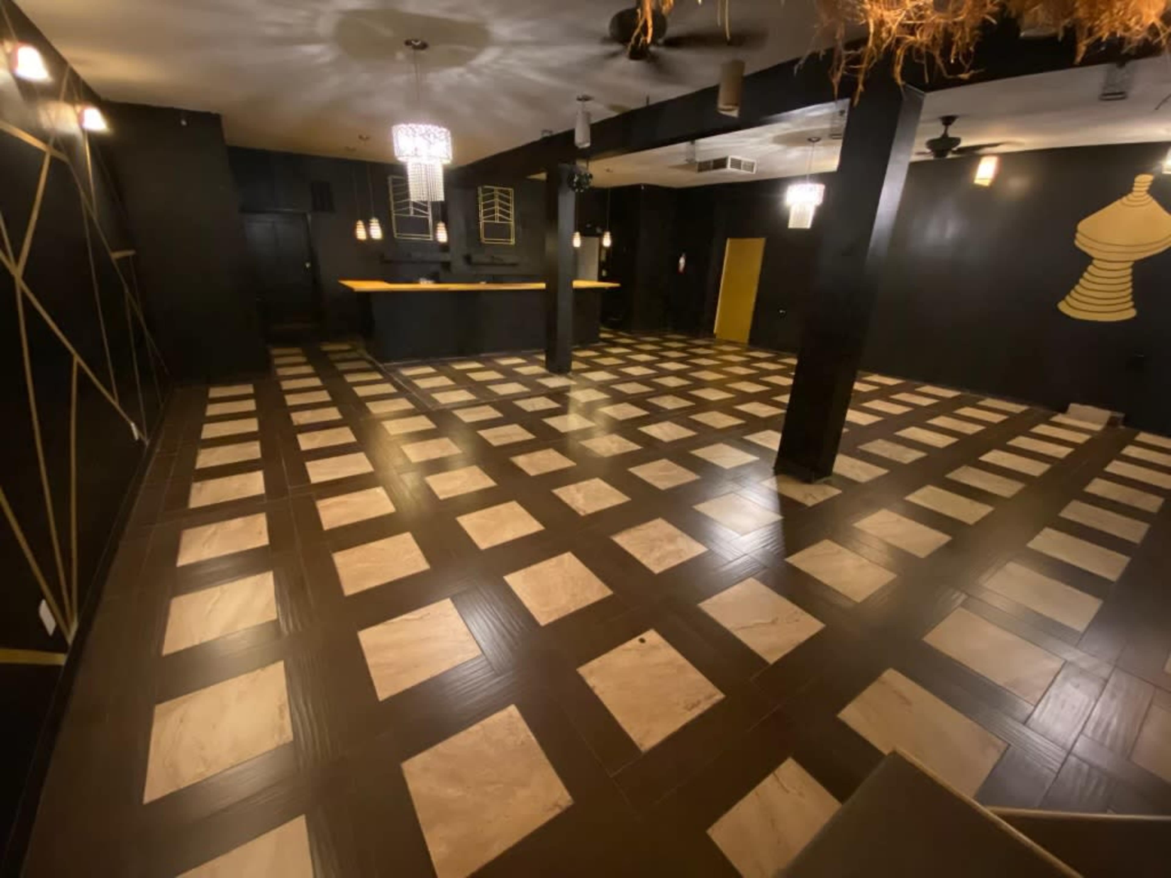 The image shows an empty interior room featuring a checkered floor pattern with alternating dark and light tiles, along with black walls and illuminated ceiling fixtures.
