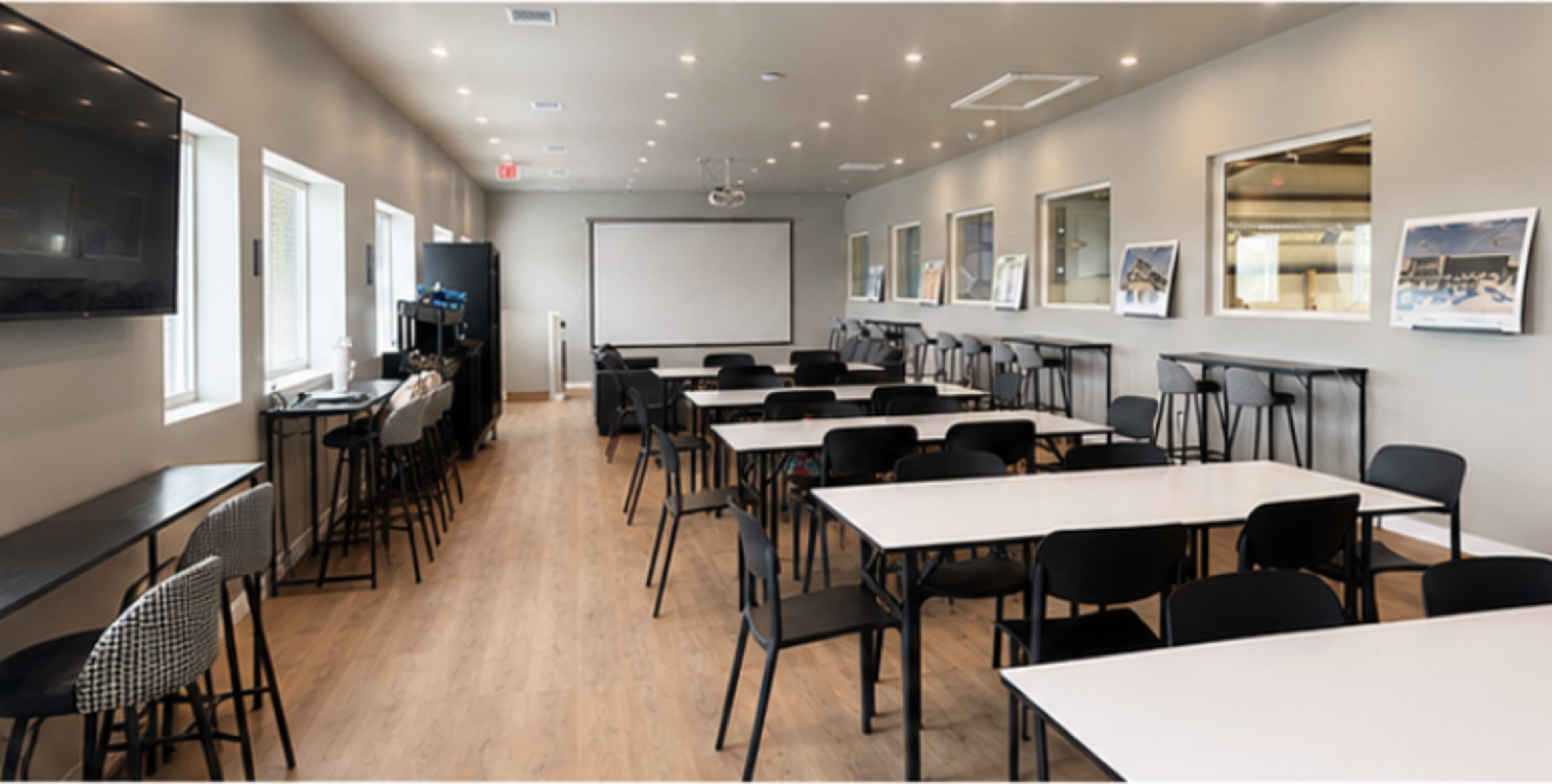 The image shows a modern conference room with rows of black chairs and tables, large windows, and a presentation screen at one end.