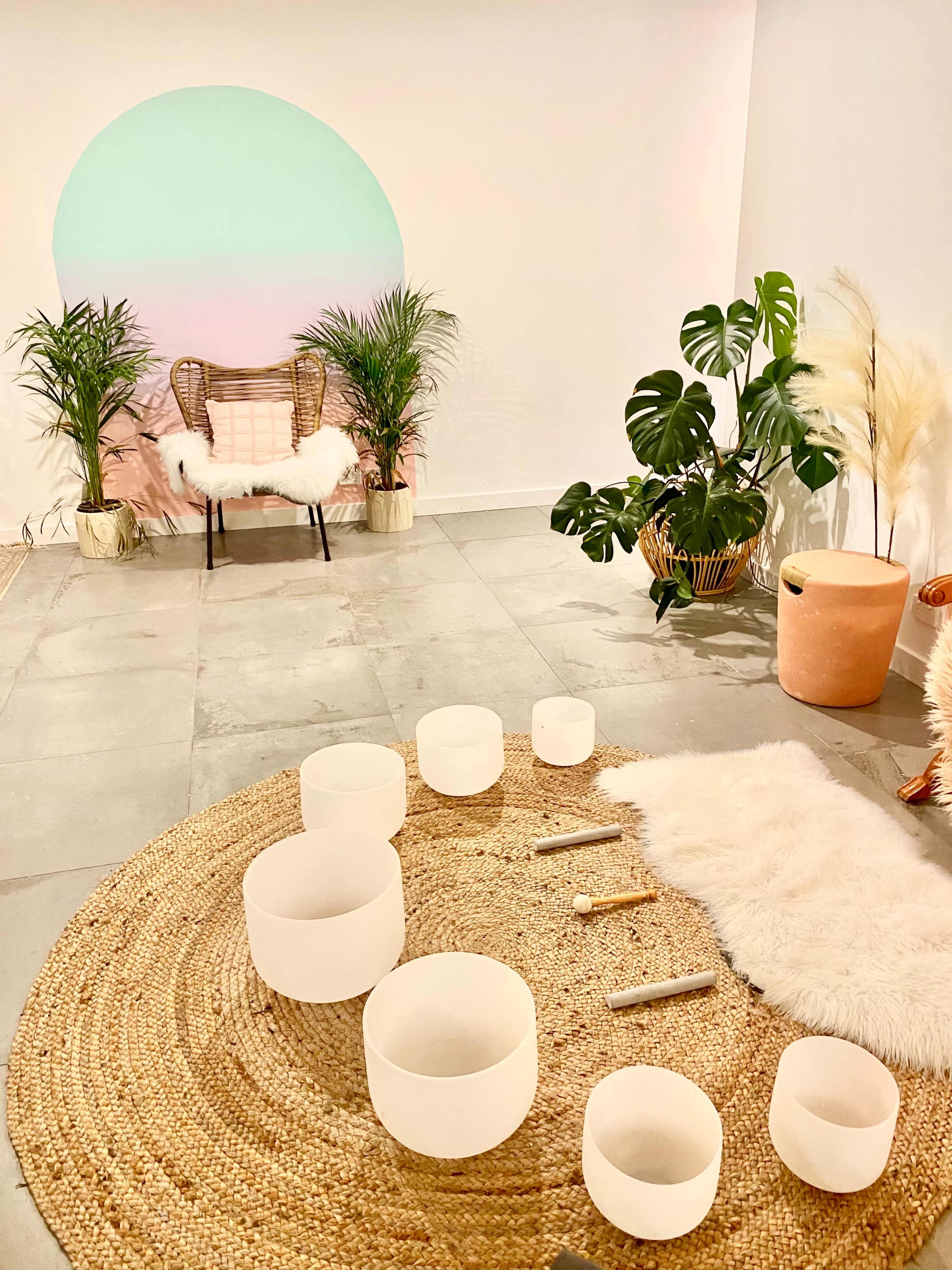 A serene indoor space featuring a round sisal rug with several crystal singing bowls arranged on it, against a backdrop of a mint and pink gradient wall with plants and a comfortable chair.