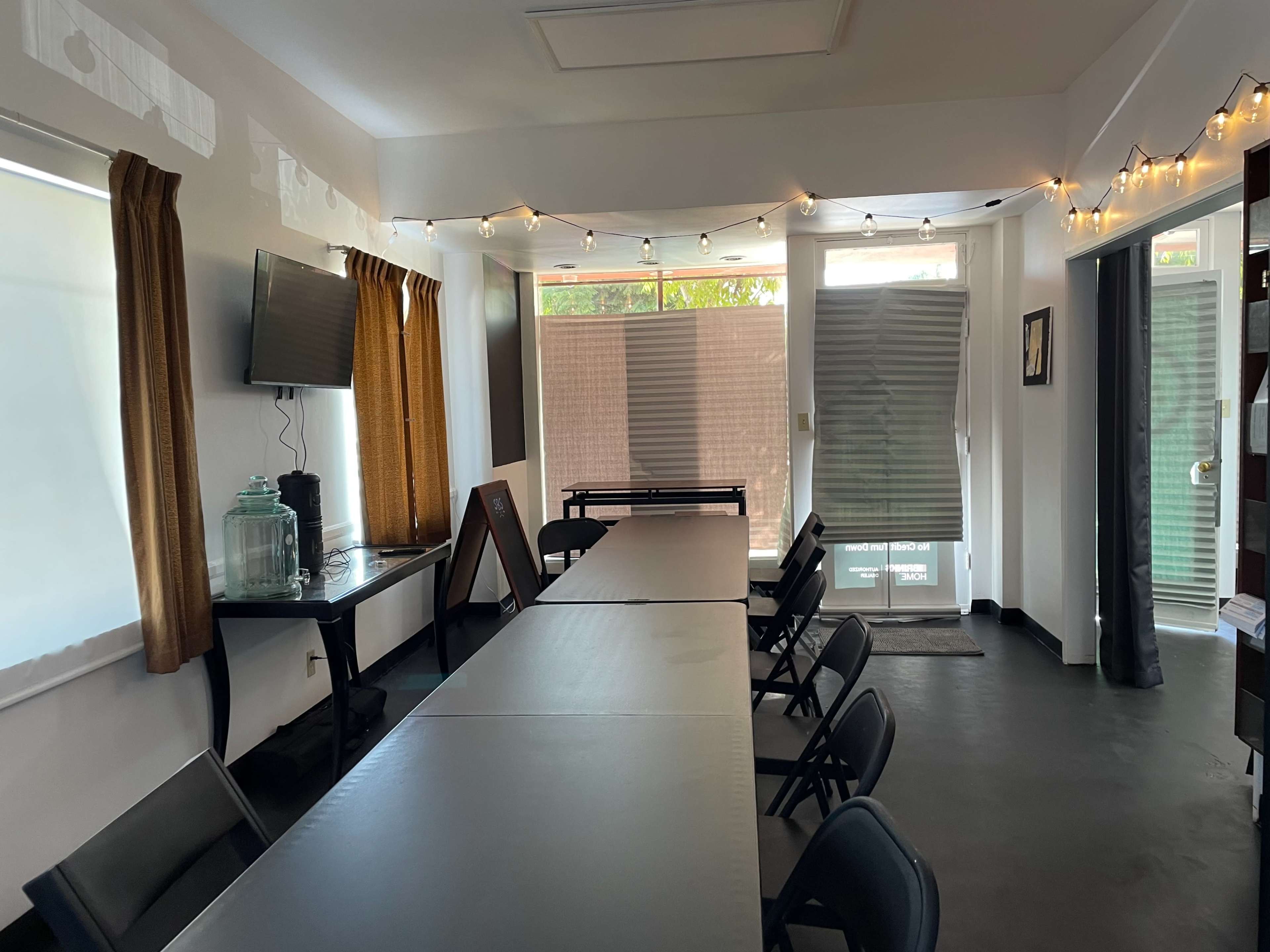 A conference room features a long table with black folding chairs, string lights along the windows, and a television mounted on the wall.