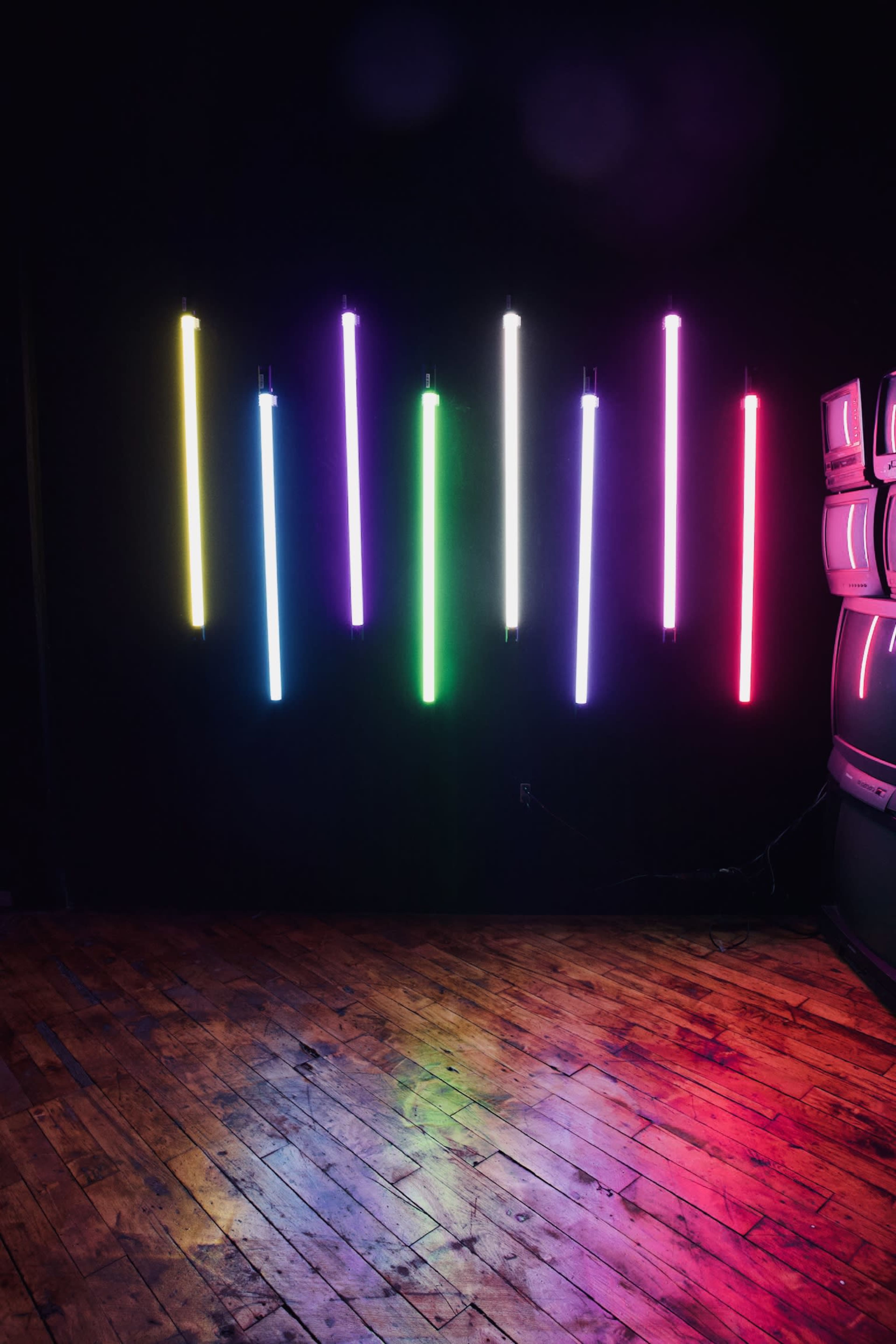 A series of vertical neon light tubes in various colors are mounted on a dark wall, reflecting off a wooden floor.