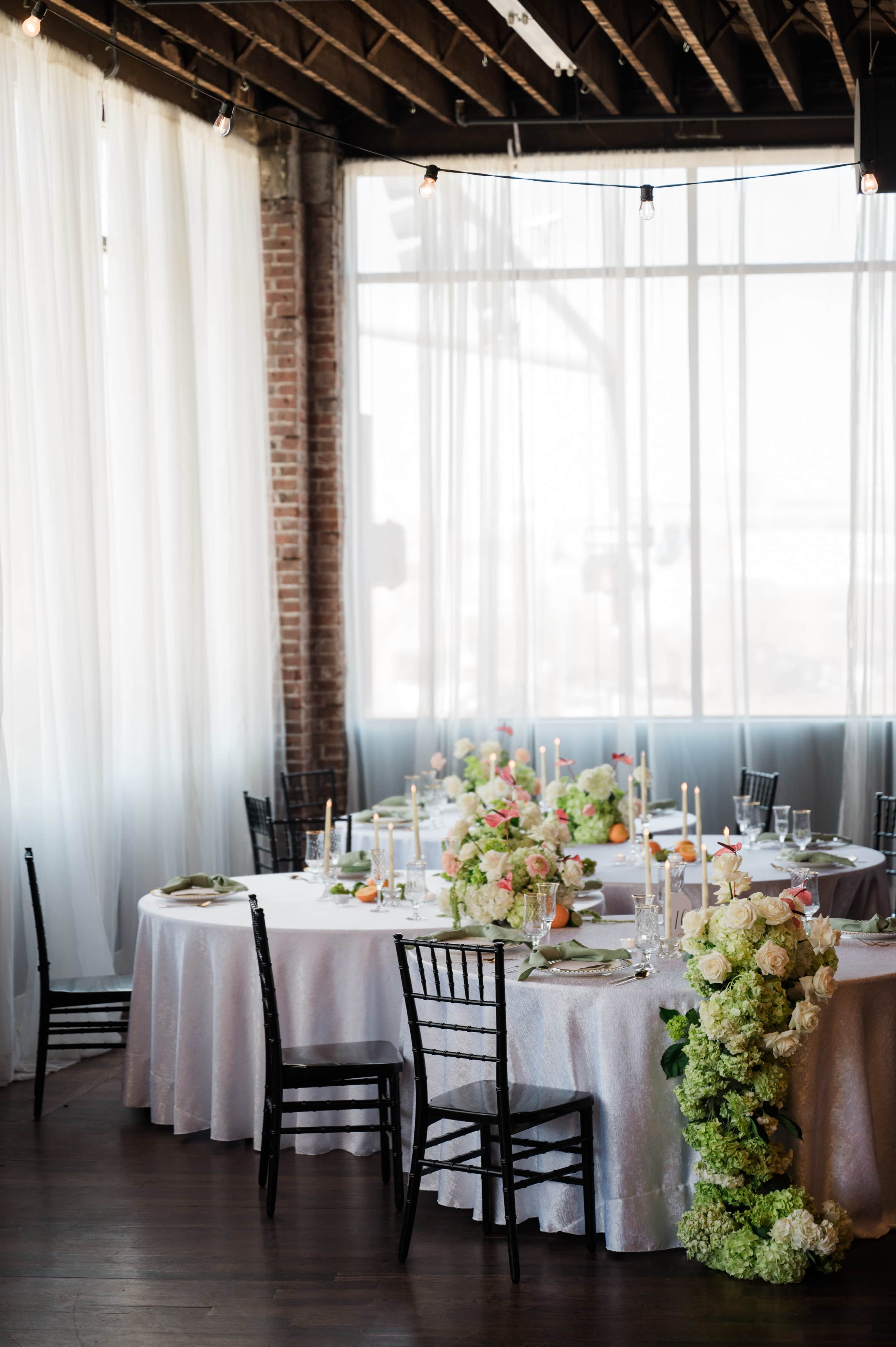 A decorated banquet hall features tables with floral centerpieces, elegant table settings, and large windows allowing natural light to fill the space.