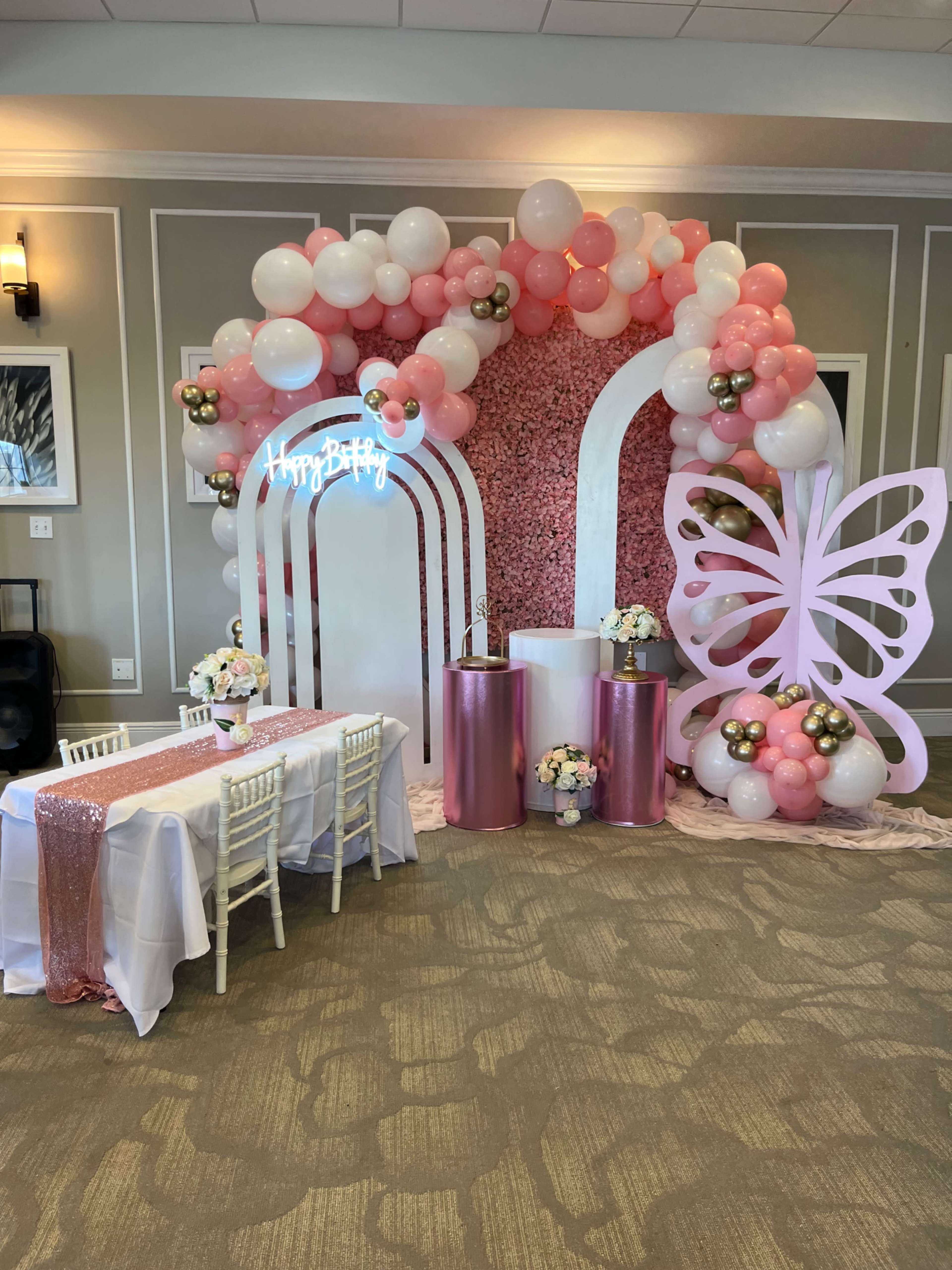 The image shows a decorative event setup featuring a balloon arch, a neon sign that says "Happy Birthday," and a table with floral arrangements, all in shades of pink and gold.