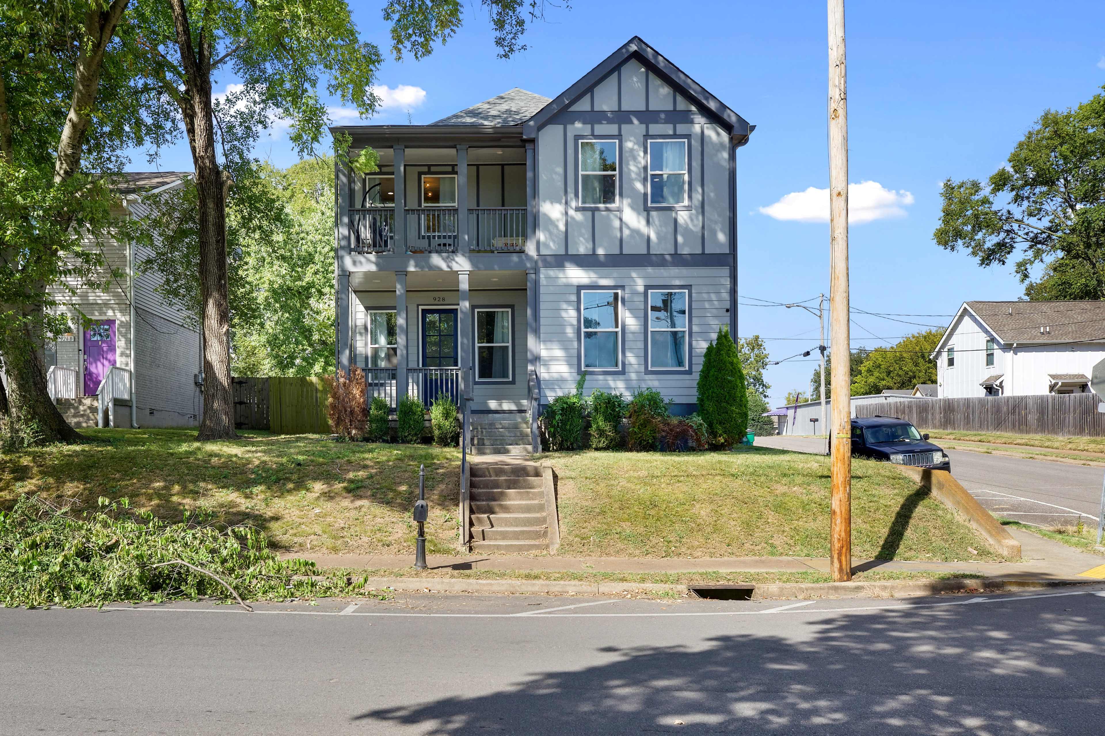 A two-story blue and gray house with a front porch is situated on a grassy lot beside a street.