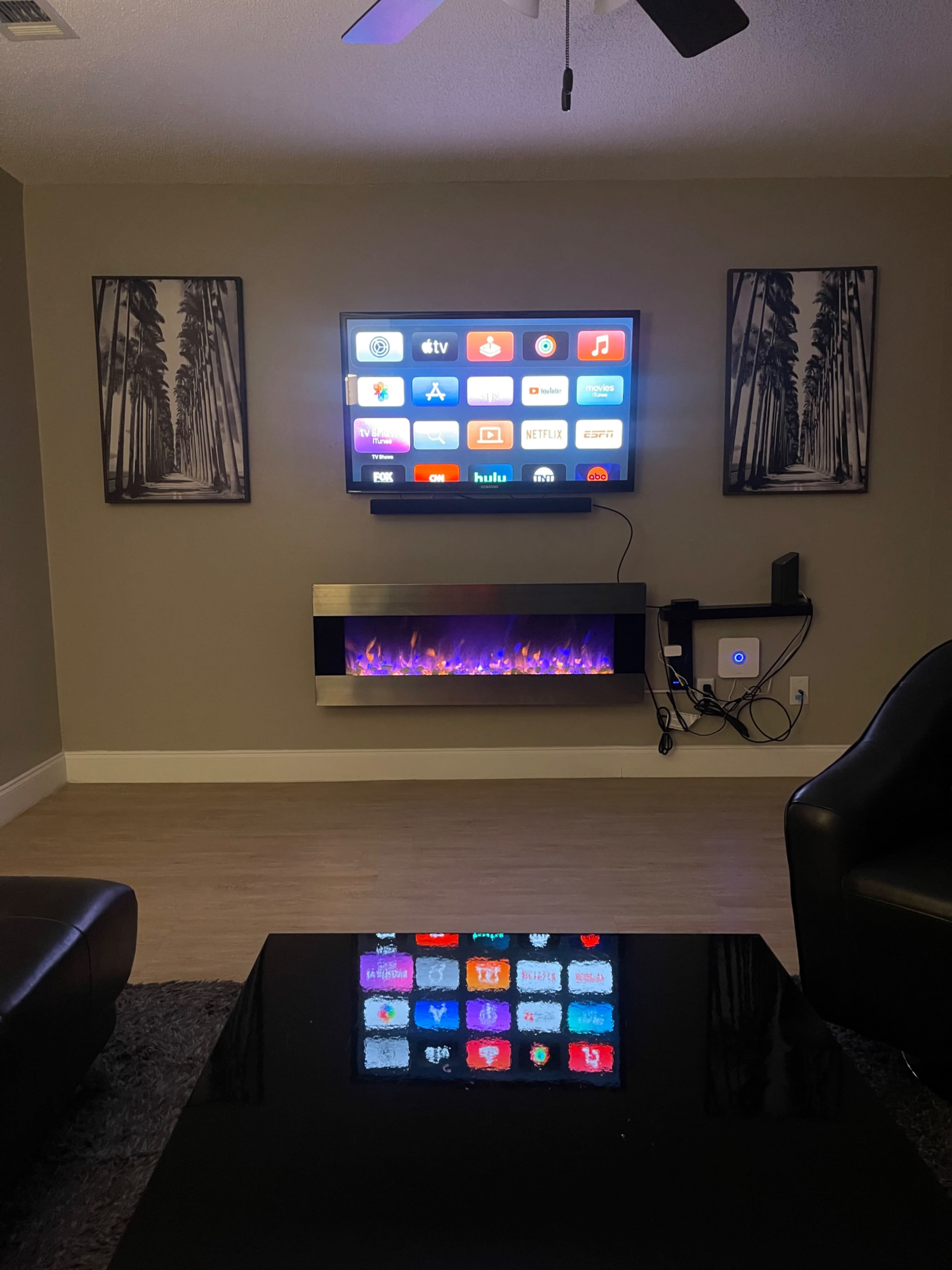 A living room with a wall-mounted TV displaying various streaming apps, above a modern electric fireplace with a colorful light feature, surrounded by two framed artworks and a black coffee table.
