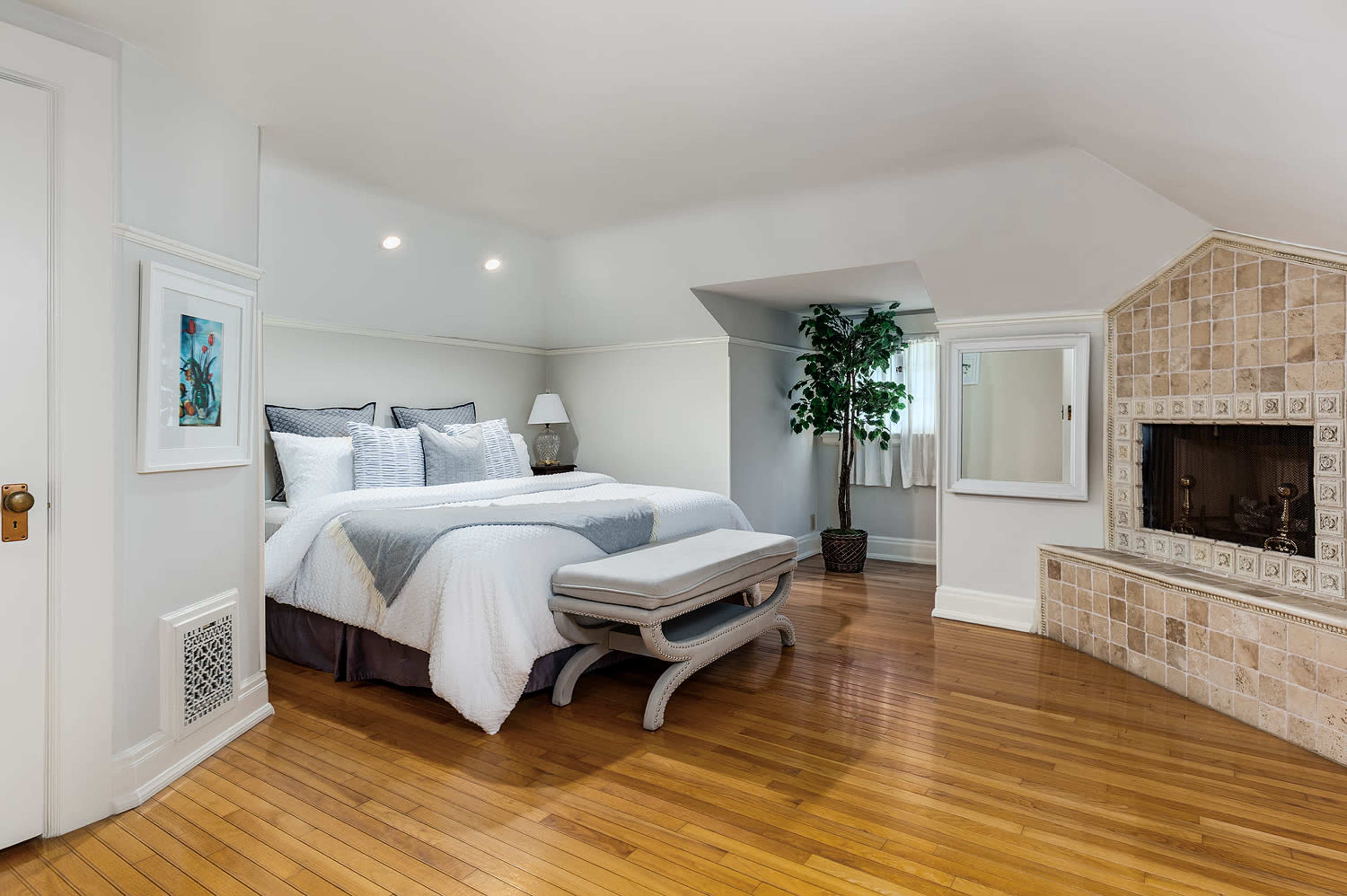 The image shows a spacious bedroom featuring a large bed with gray bedding, a wooden floor, and a fireplace with tiled accents.
