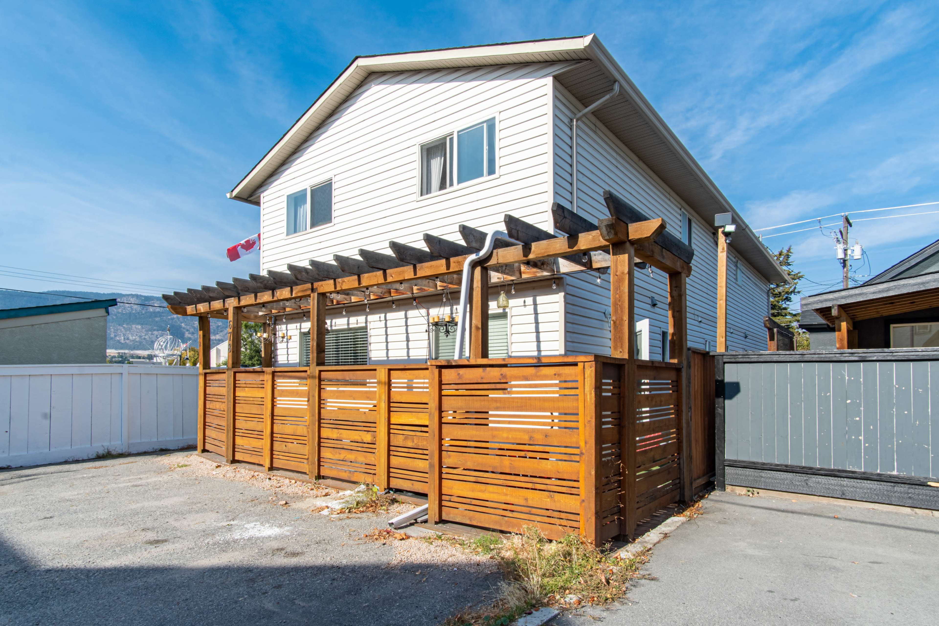 A two-story house with white siding and a wooden fence, featuring a pergola in front, set against a clear blue sky.