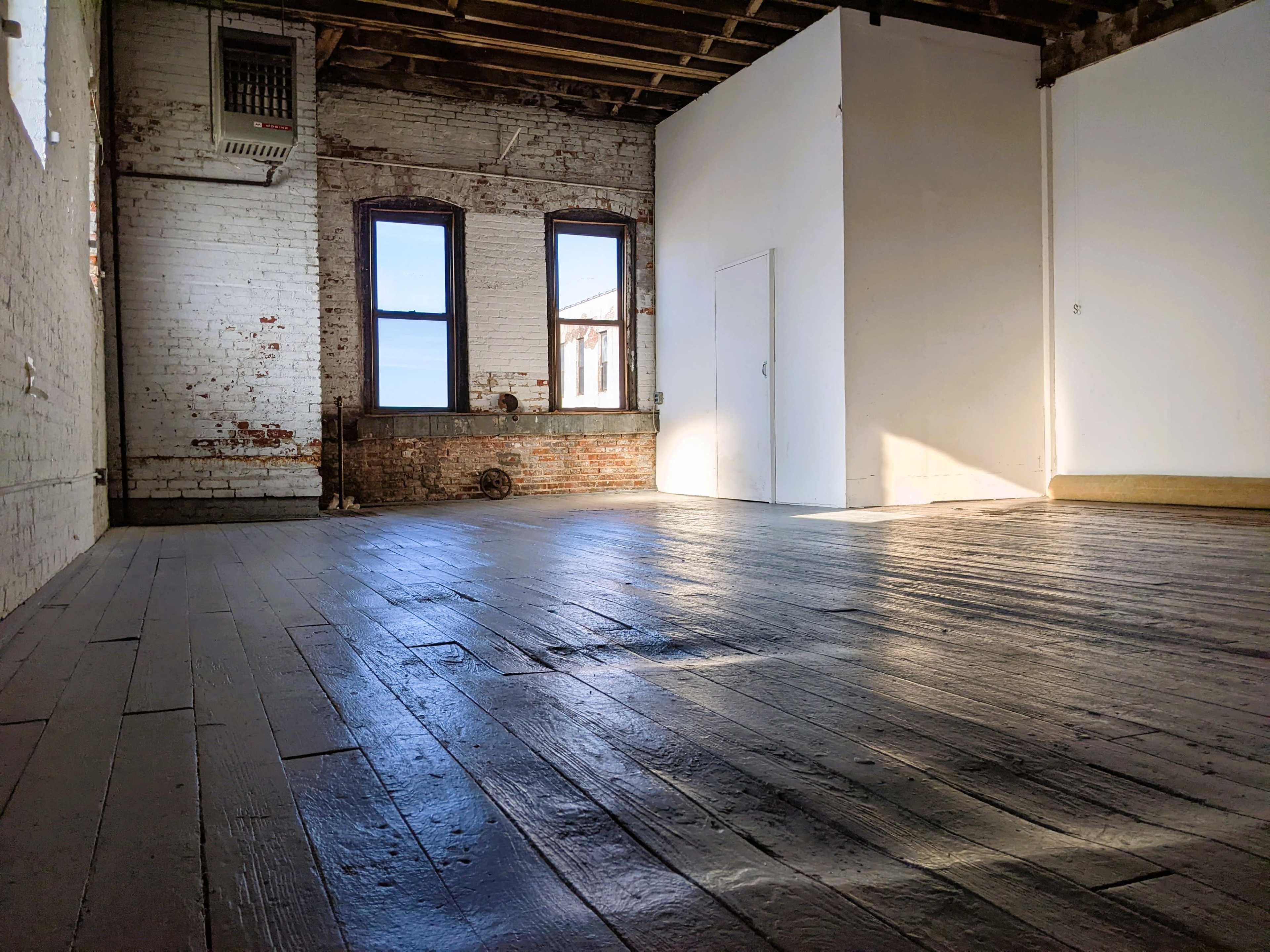 An empty room with wooden floors, exposed brick walls, and large windows letting in light.