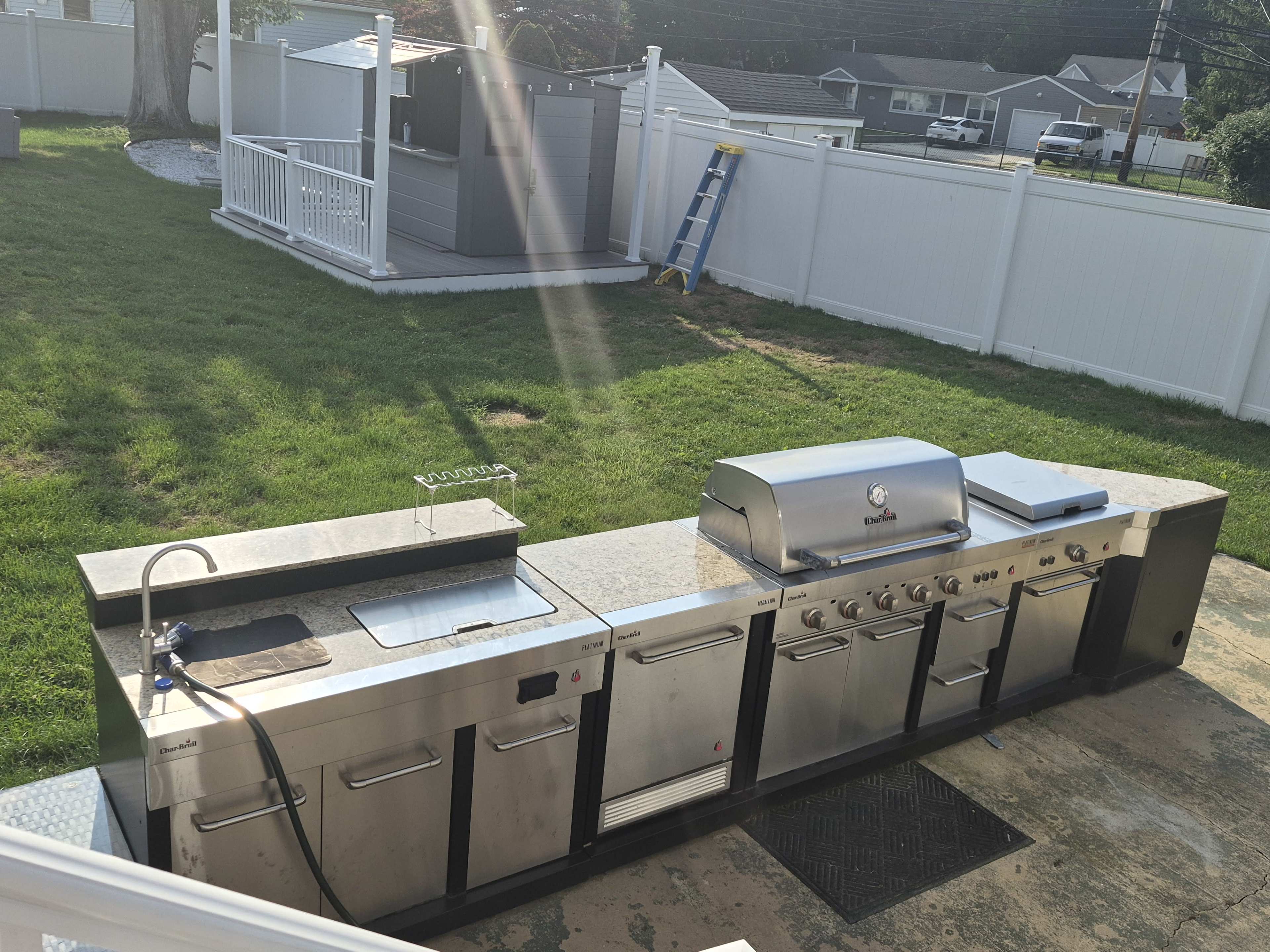 The image shows an outdoor kitchen setup featuring a grill, sink, and countertop in a grassy backyard area.