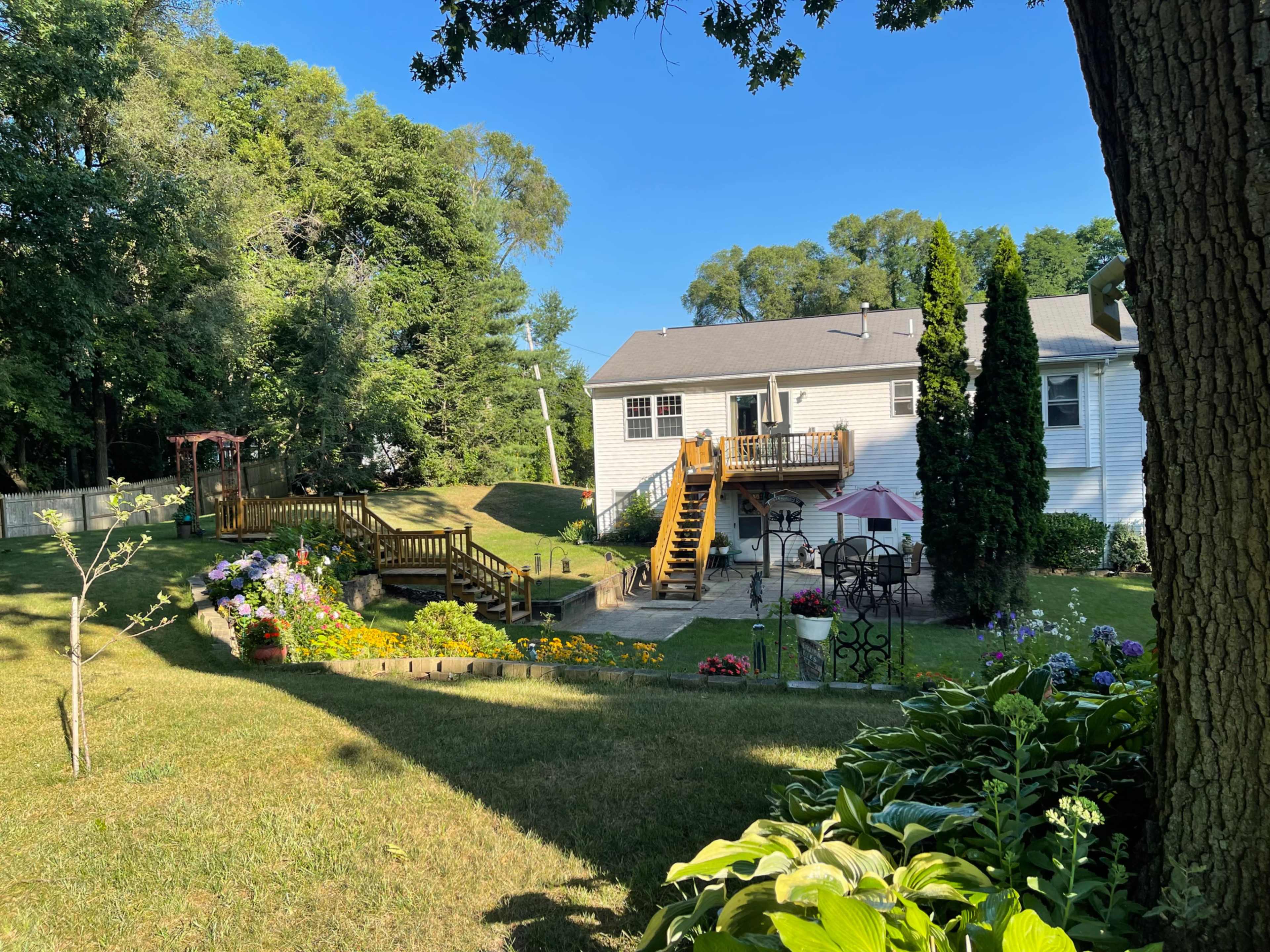 A well-maintained backyard features a white house with a deck, surrounded by landscaped gardens and trees.