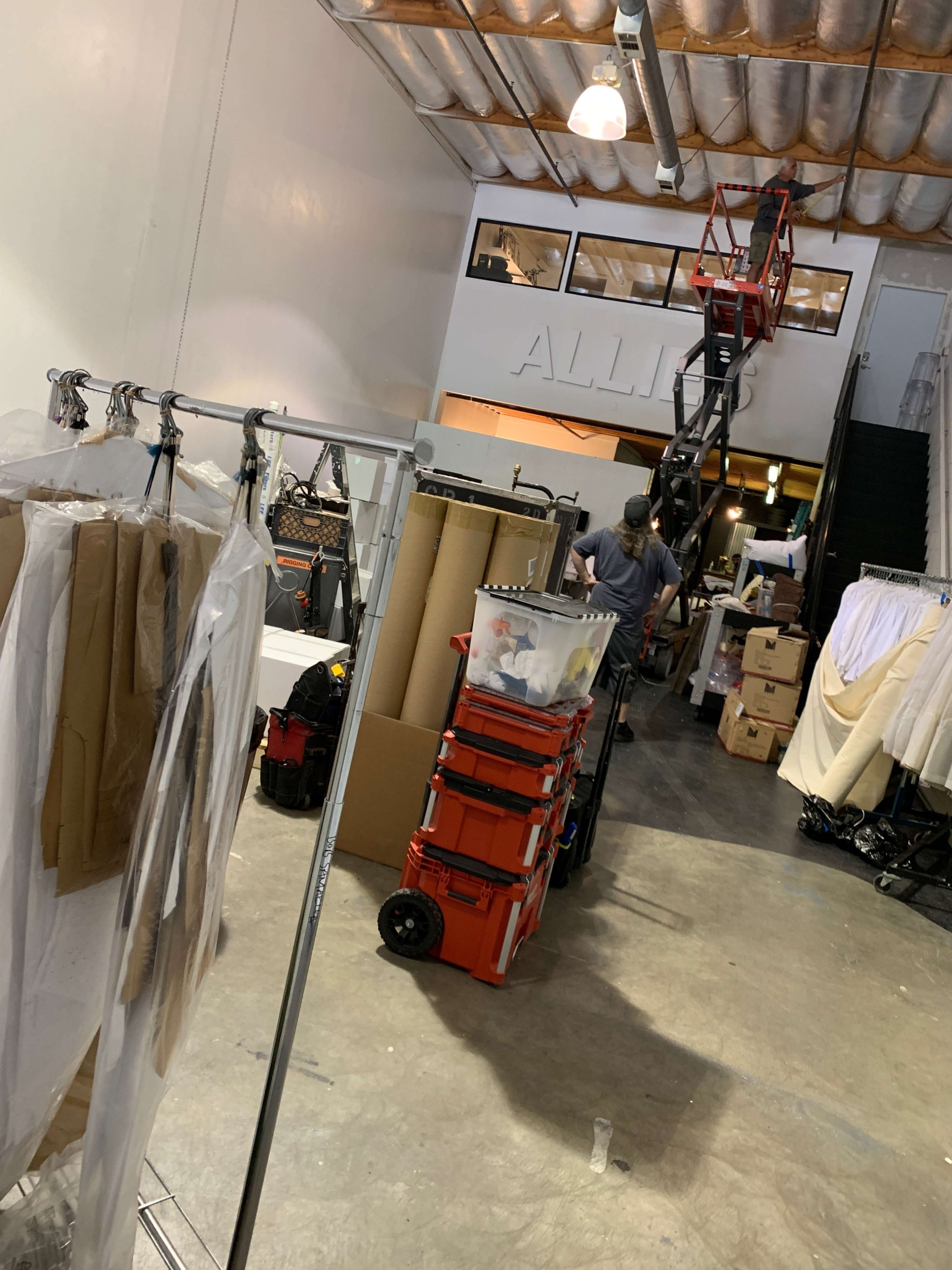 The image shows an industrial workspace with clothing racks on the left, storage boxes, and a lift being operated by a person ascending a staircase in the background.